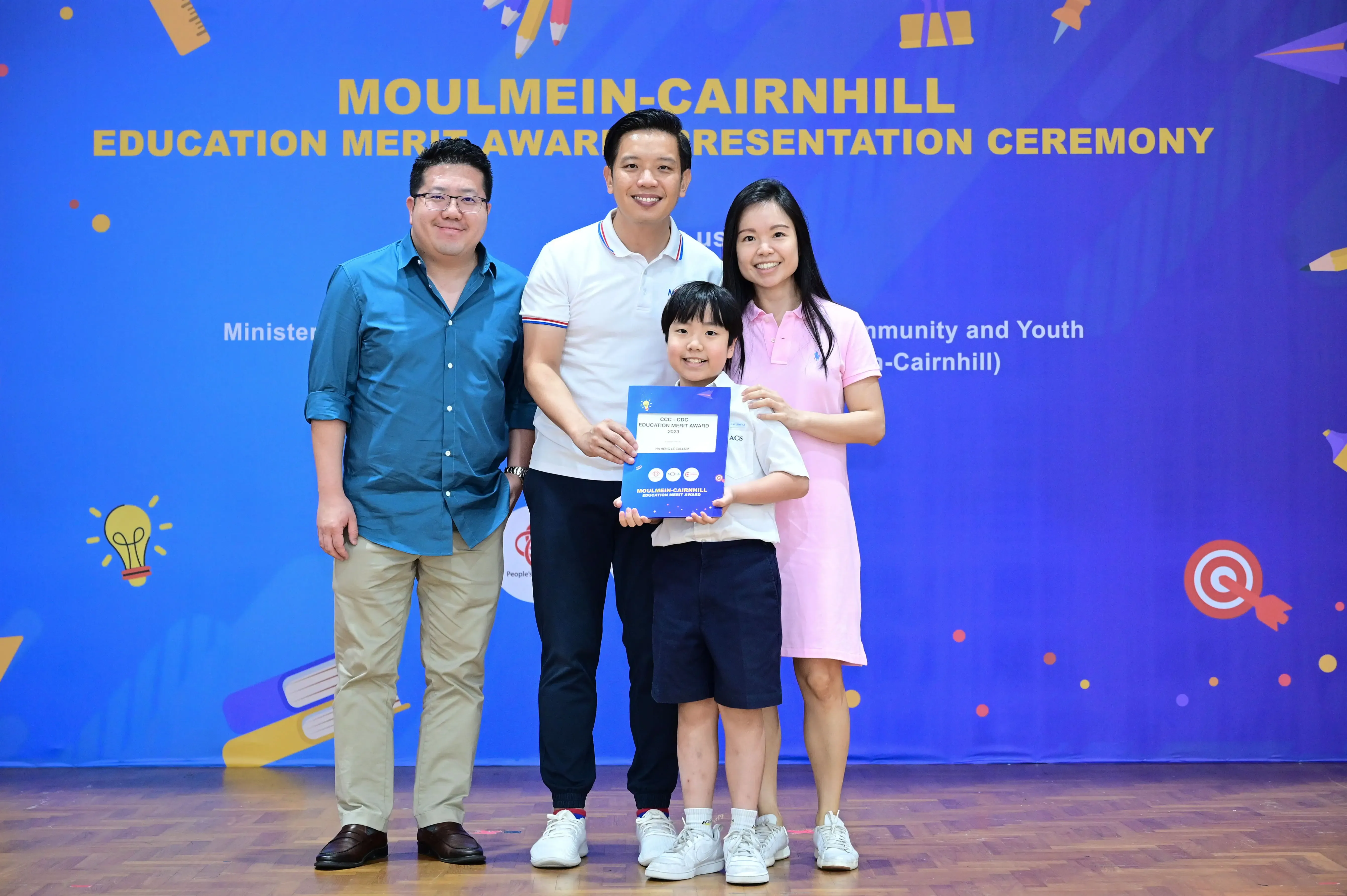 Four people pose smiling at the Moulmein-Cairnhill Education Merit Award Presentation Ceremony, a child holding a blue award certificate.