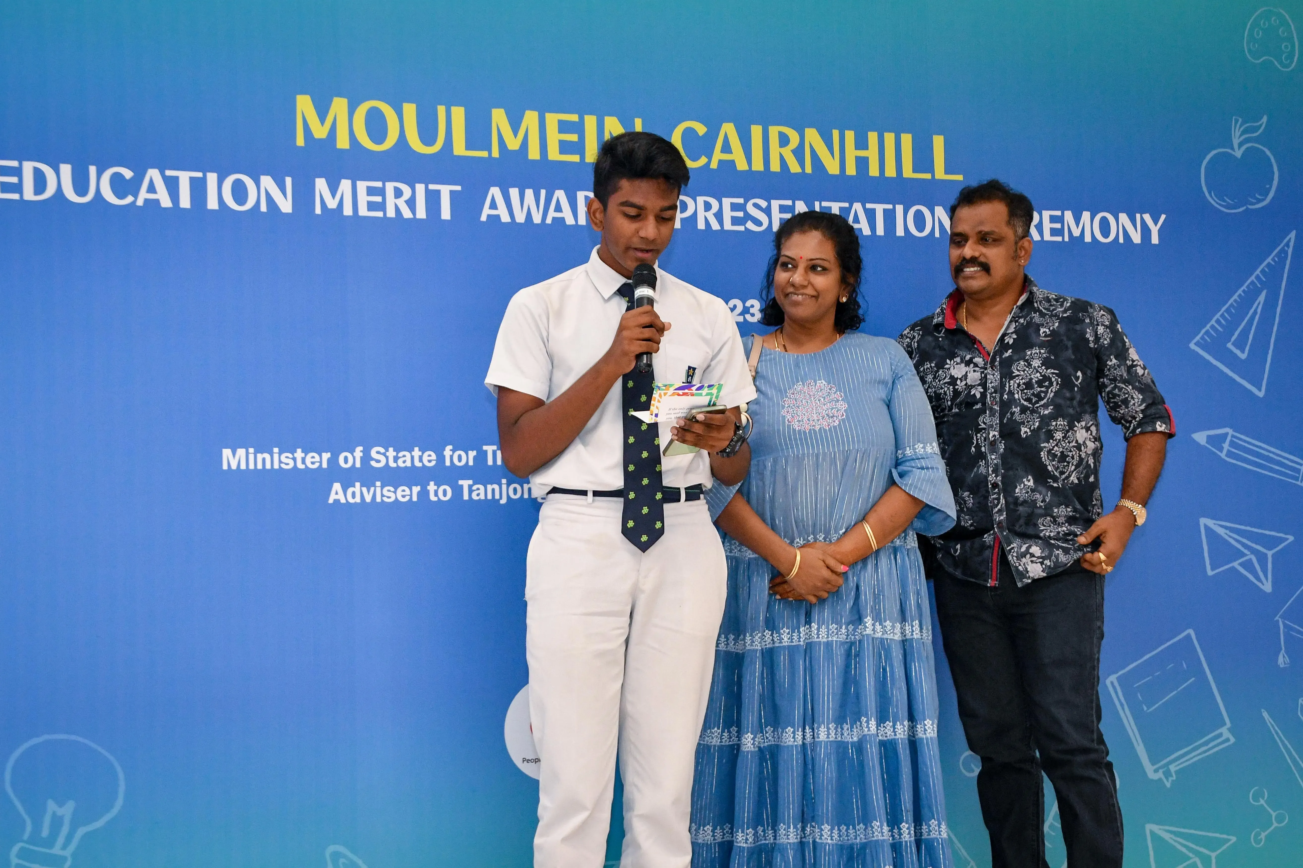 A teenage boy in white uniform speaks into a microphone at Moulmein Cairnhill Education Merit Award Ceremony, flanked by two adults.