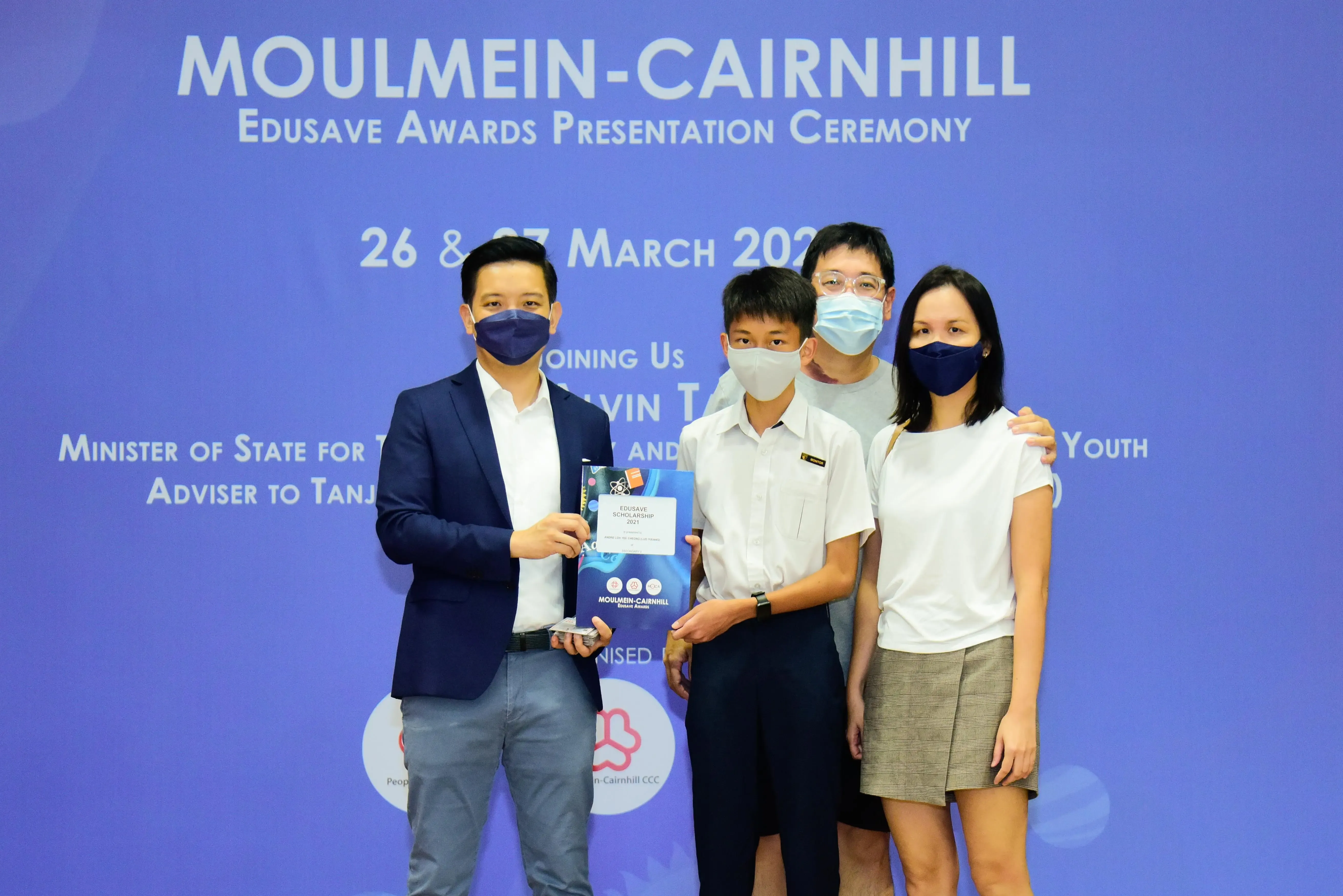 Four masked people pose at the Moulmein-Cairnhill Edusave Awards Presentation Ceremony, holding an award booklet.