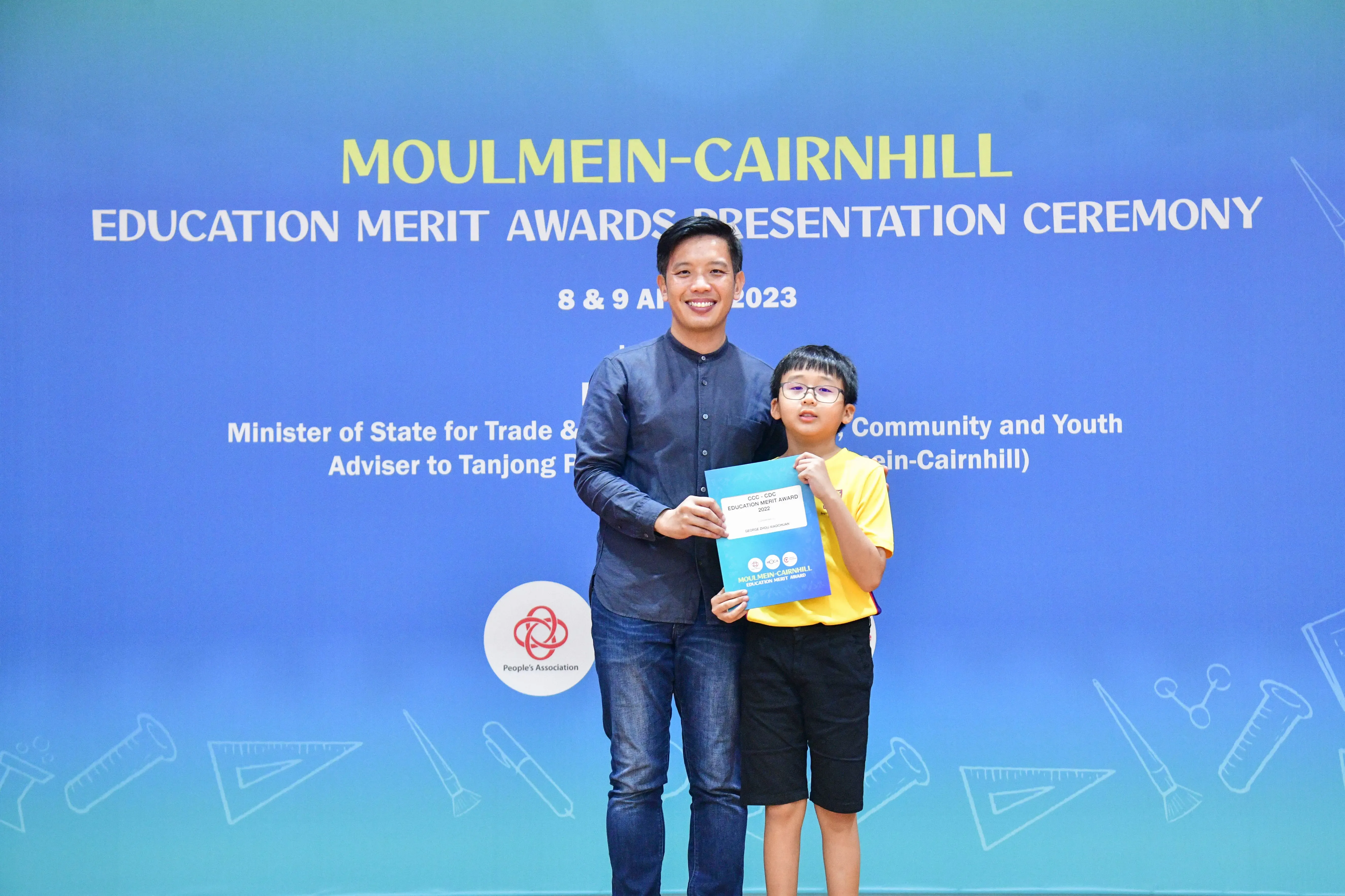 An adult and a young boy holding a certificate stand before a Moulmein-Cairnhill Education Merit Awards banner.