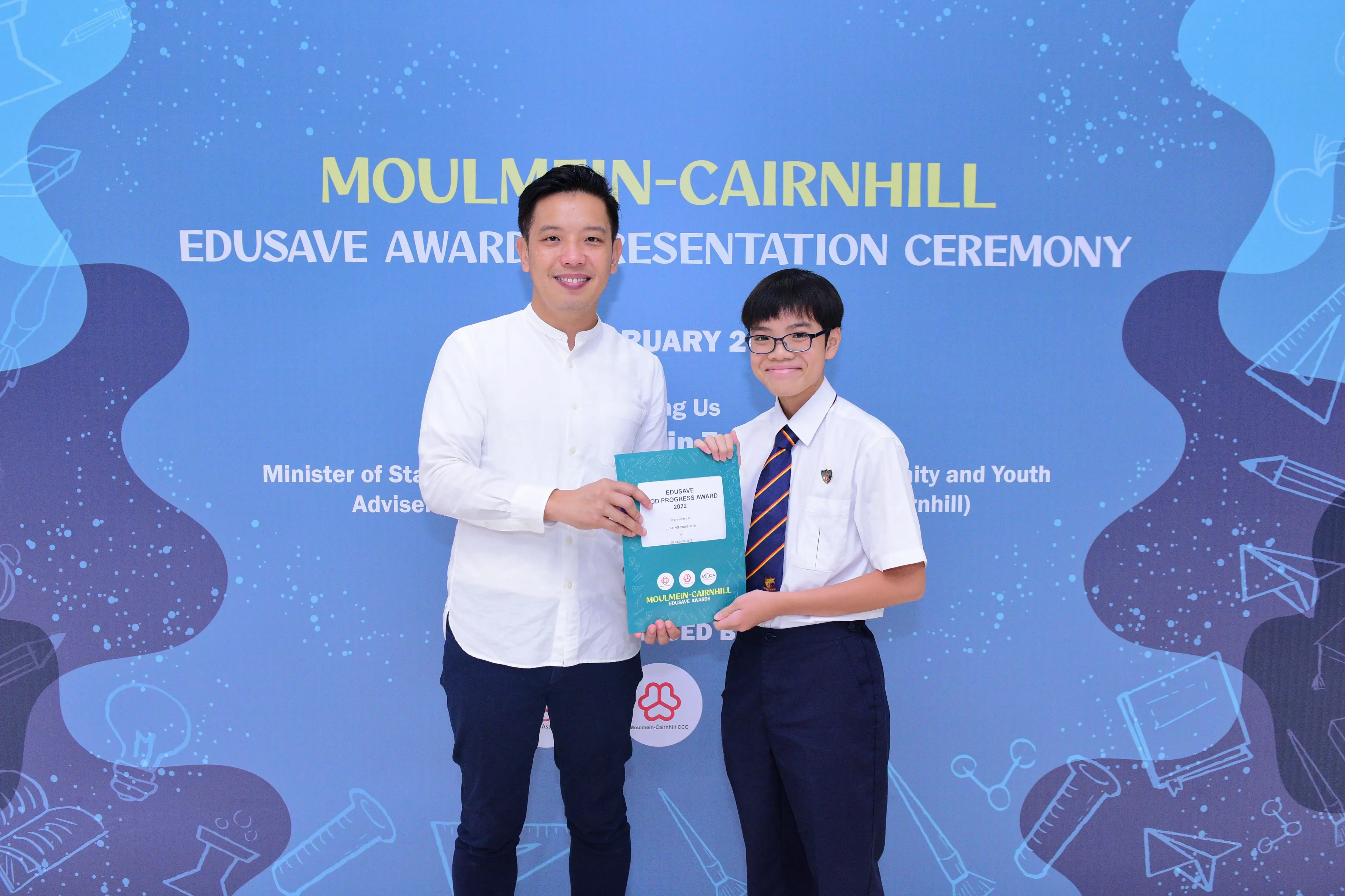 Two people hold a certificate together in front of a Moulmein-Cairnhill Edusave Award Presentation Ceremony banner.