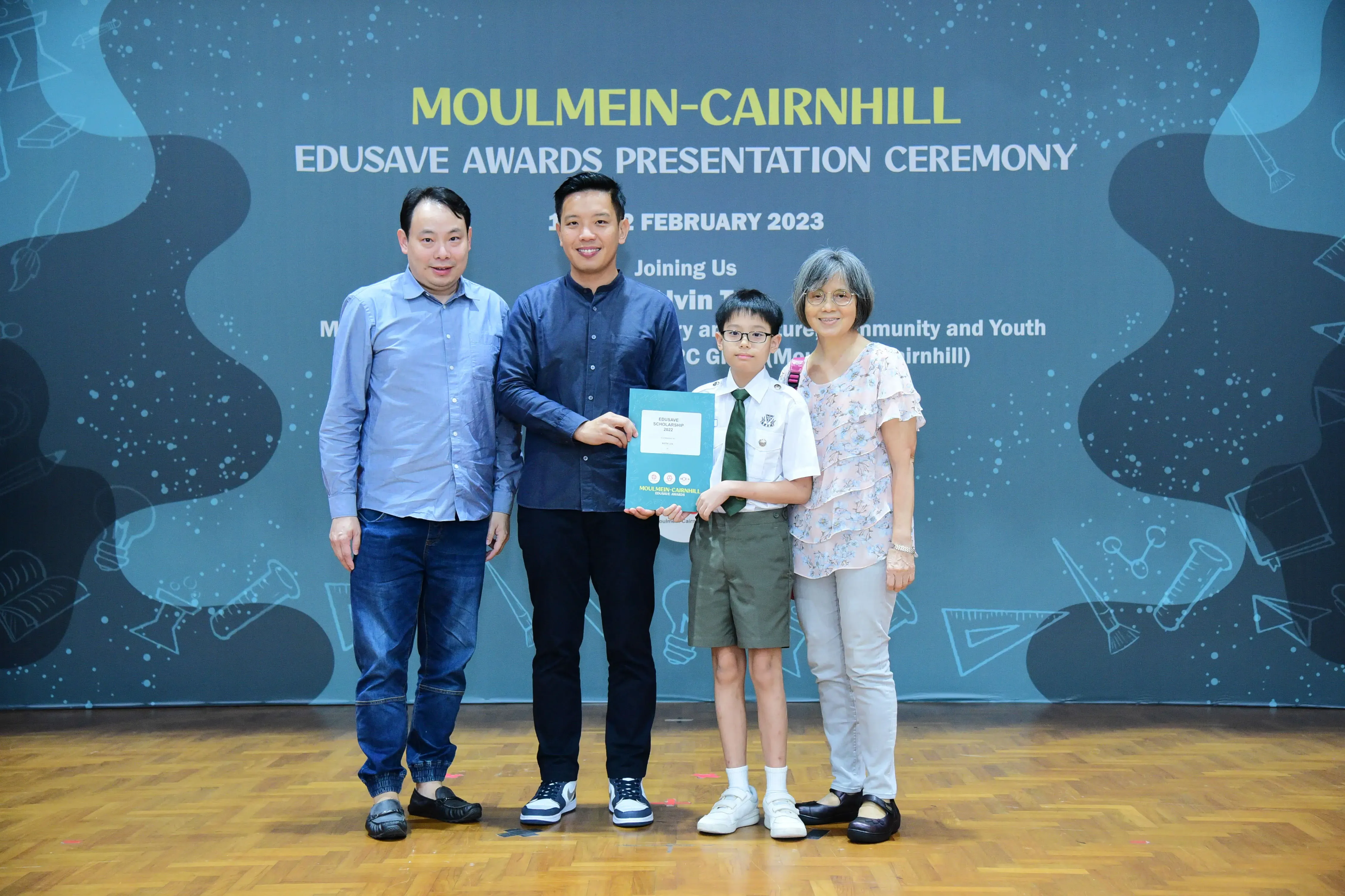 Four people pose at the Moulmein-Cairnhill Edusave Awards Ceremony, with a boy in school uniform holding a certificate.