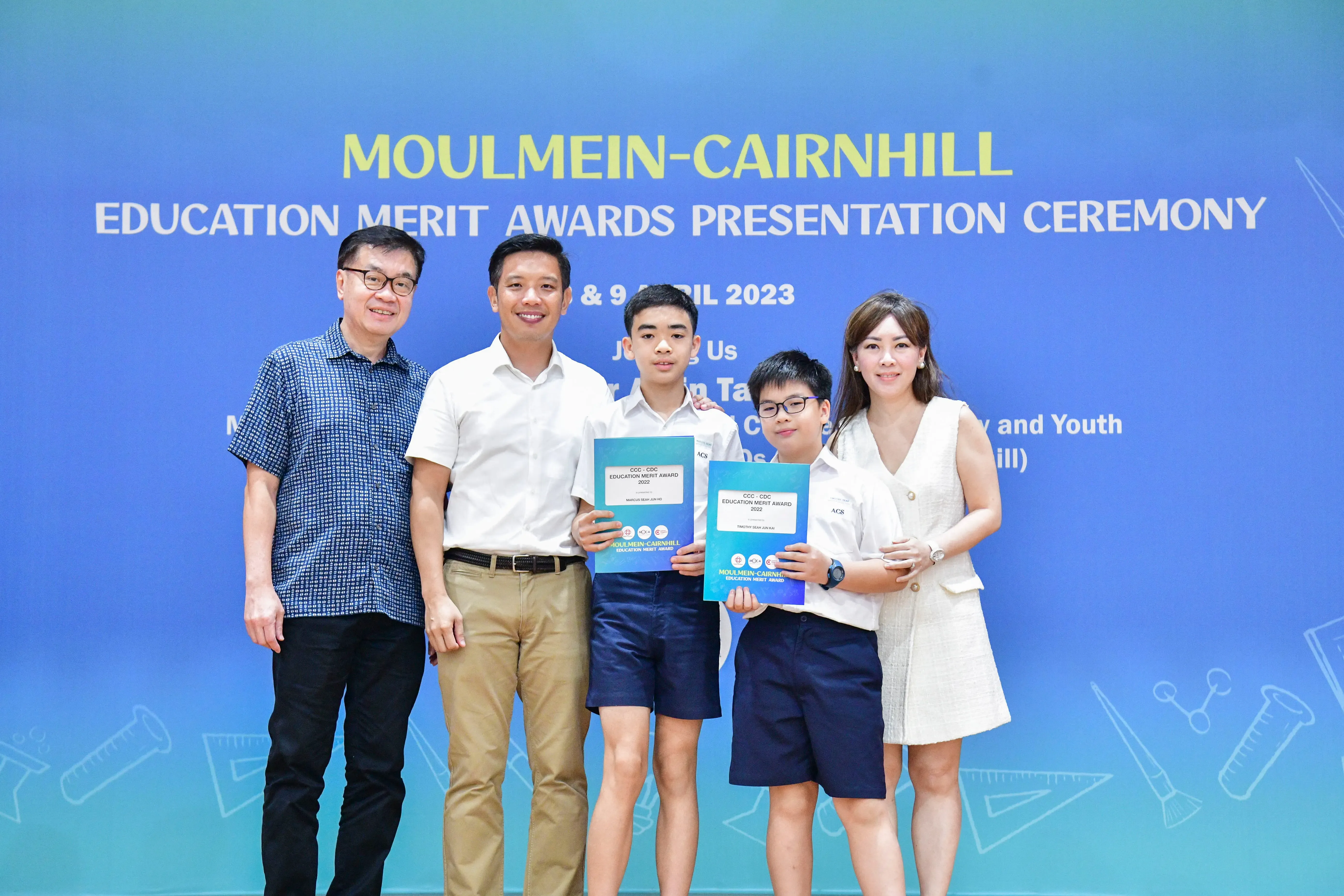 Five people pose before a Moulmein-Cairnhill Education Merit Awards 2023 banner, two boys holding award certificates.