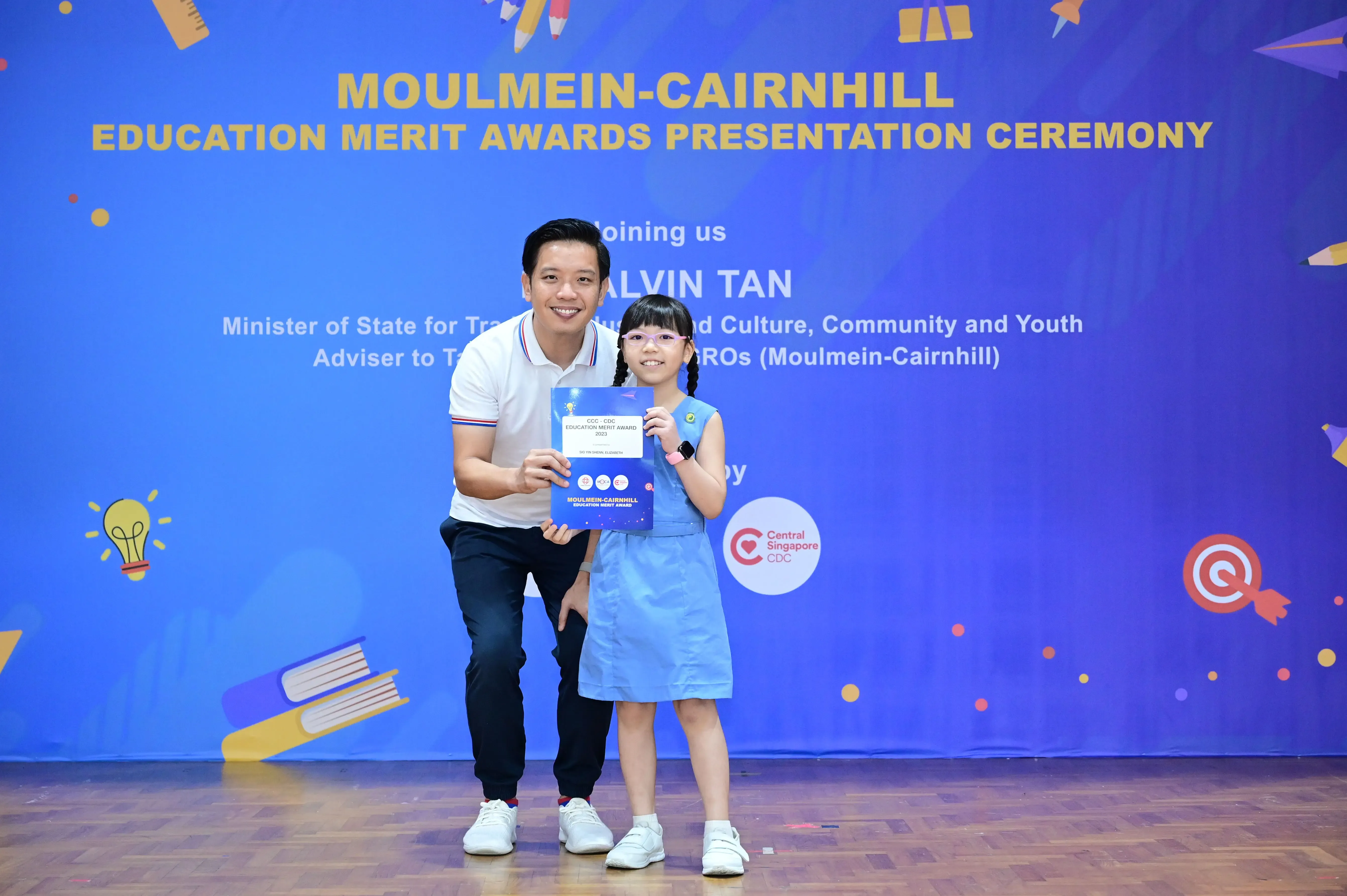 A man and a young girl hold an award certificate at the Moulmein-Cairnhill Education Merit Awards ceremony.