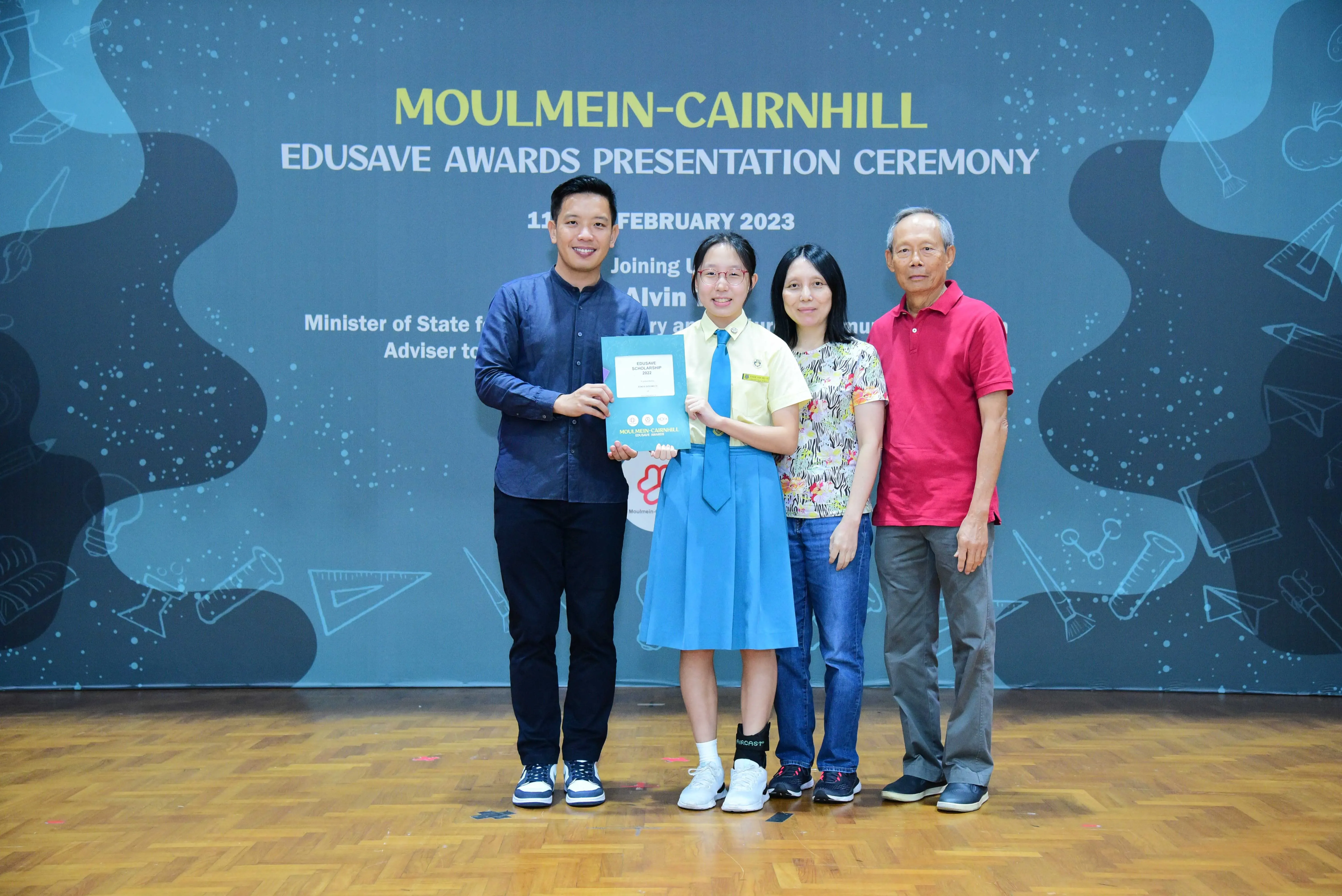 Four people pose on a stage at the Moulmein-Cairnhill Edusave Awards Presentation Ceremony, student holding a certificate.