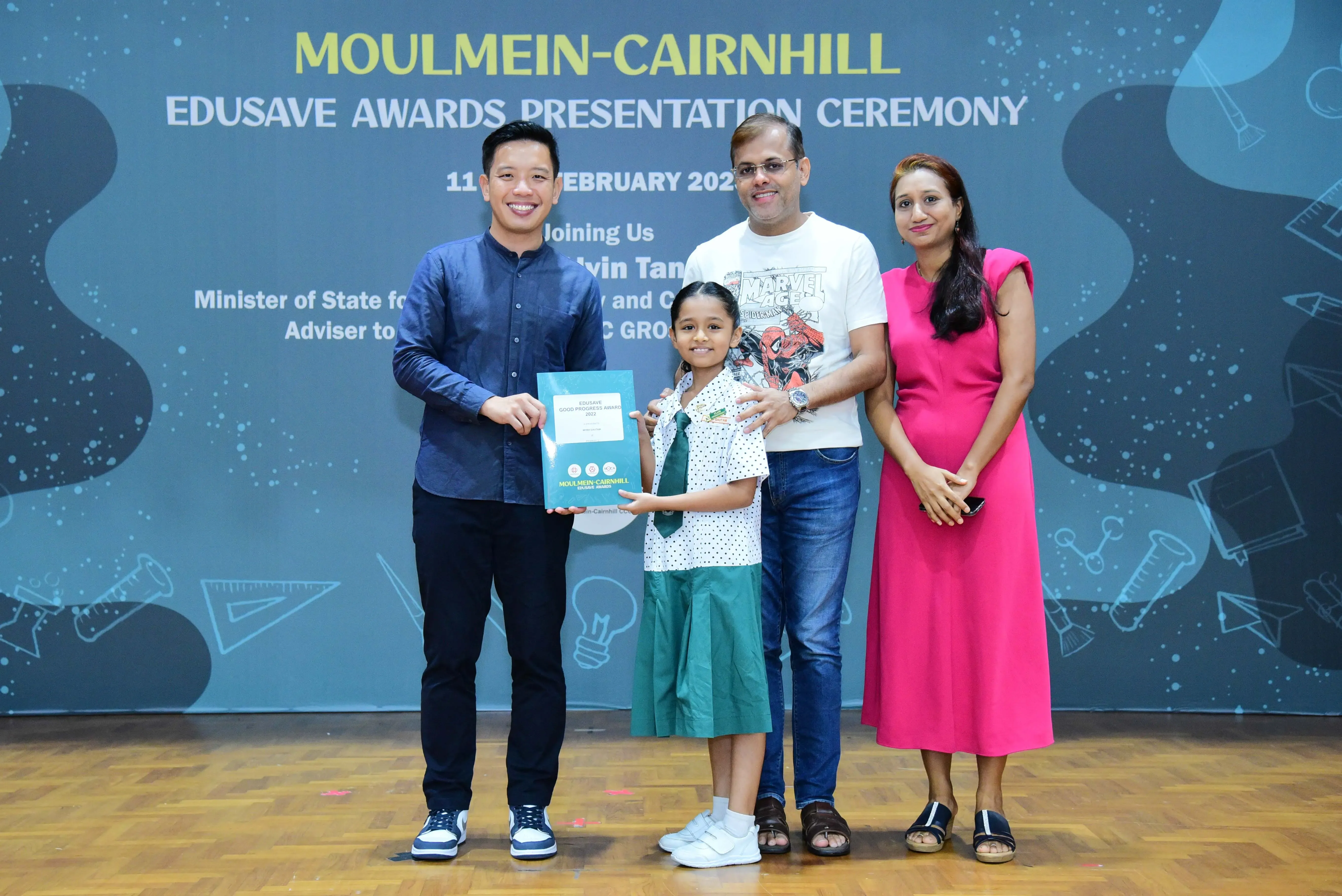 Four people pose at the Moulmein-Cairnhill Edusave Awards Presentation Ceremony, with a young girl holding an award.