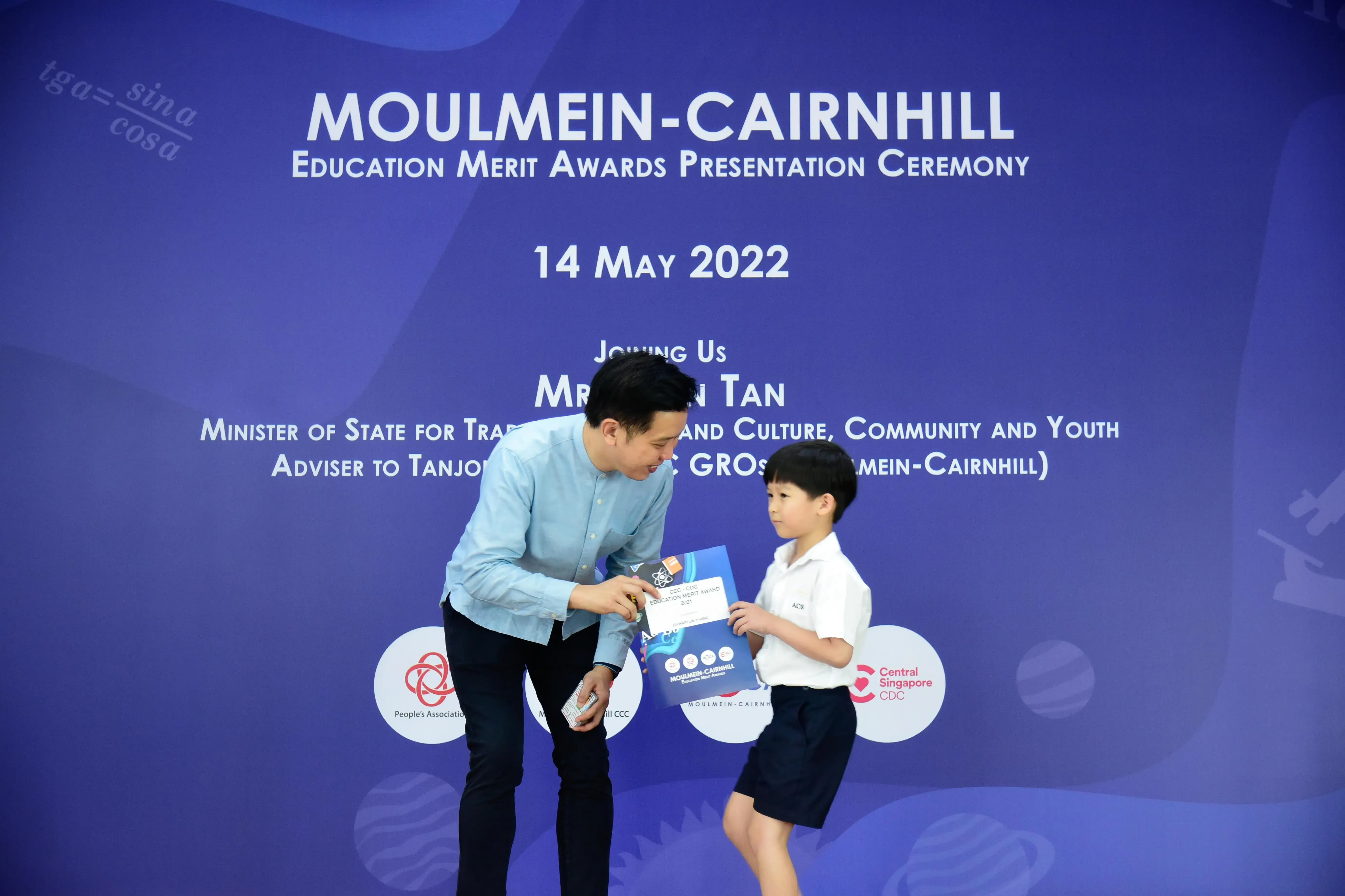 A man in a blue shirt hands a certificate to a young boy in school uniform at the Moulmein-Cairnhill Education Merit Awards 2022.