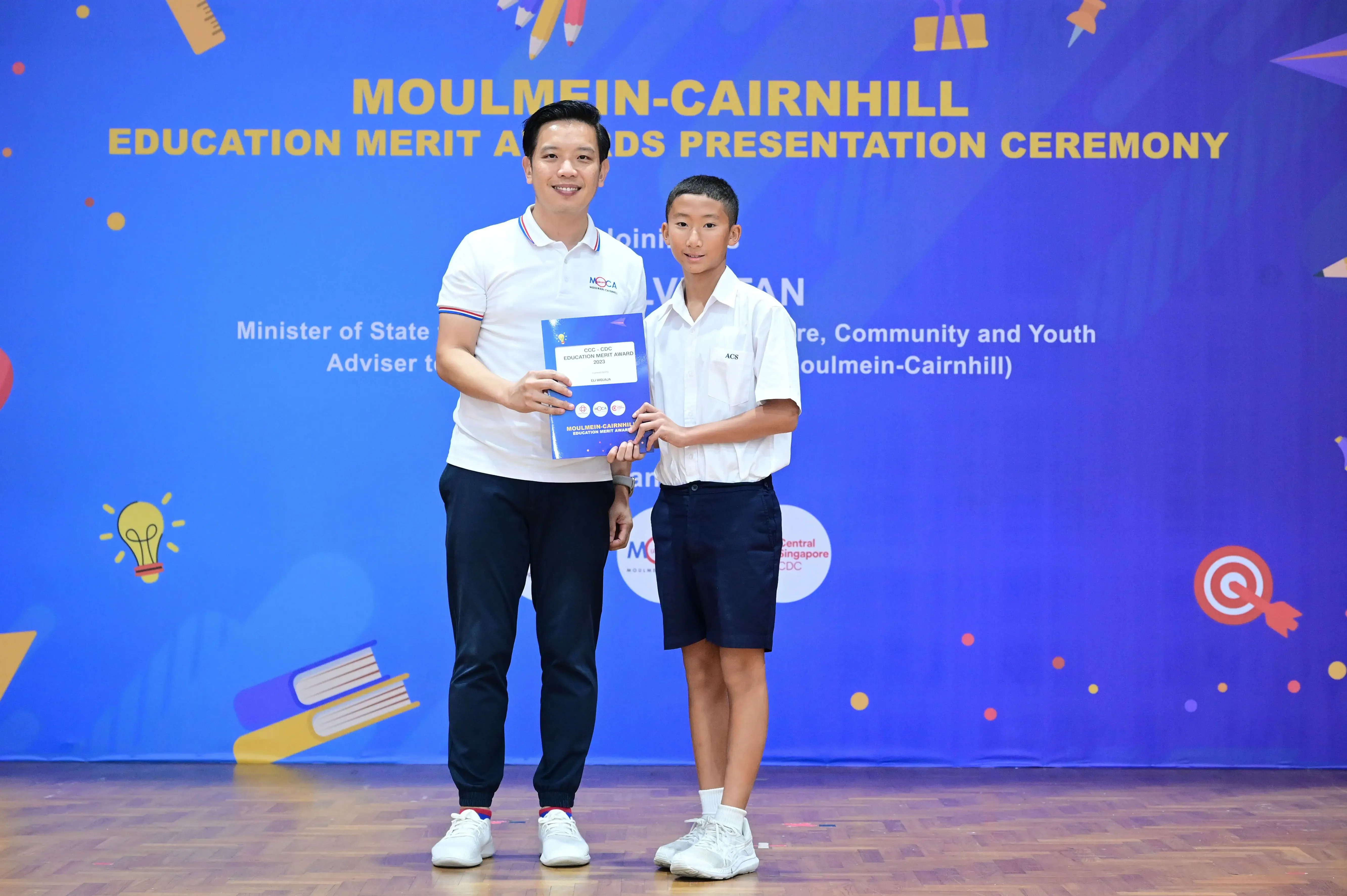 An adult and a young student hold an award booklet on stage at the Moulmein-Cairnhill Education Merit Awards ceremony.