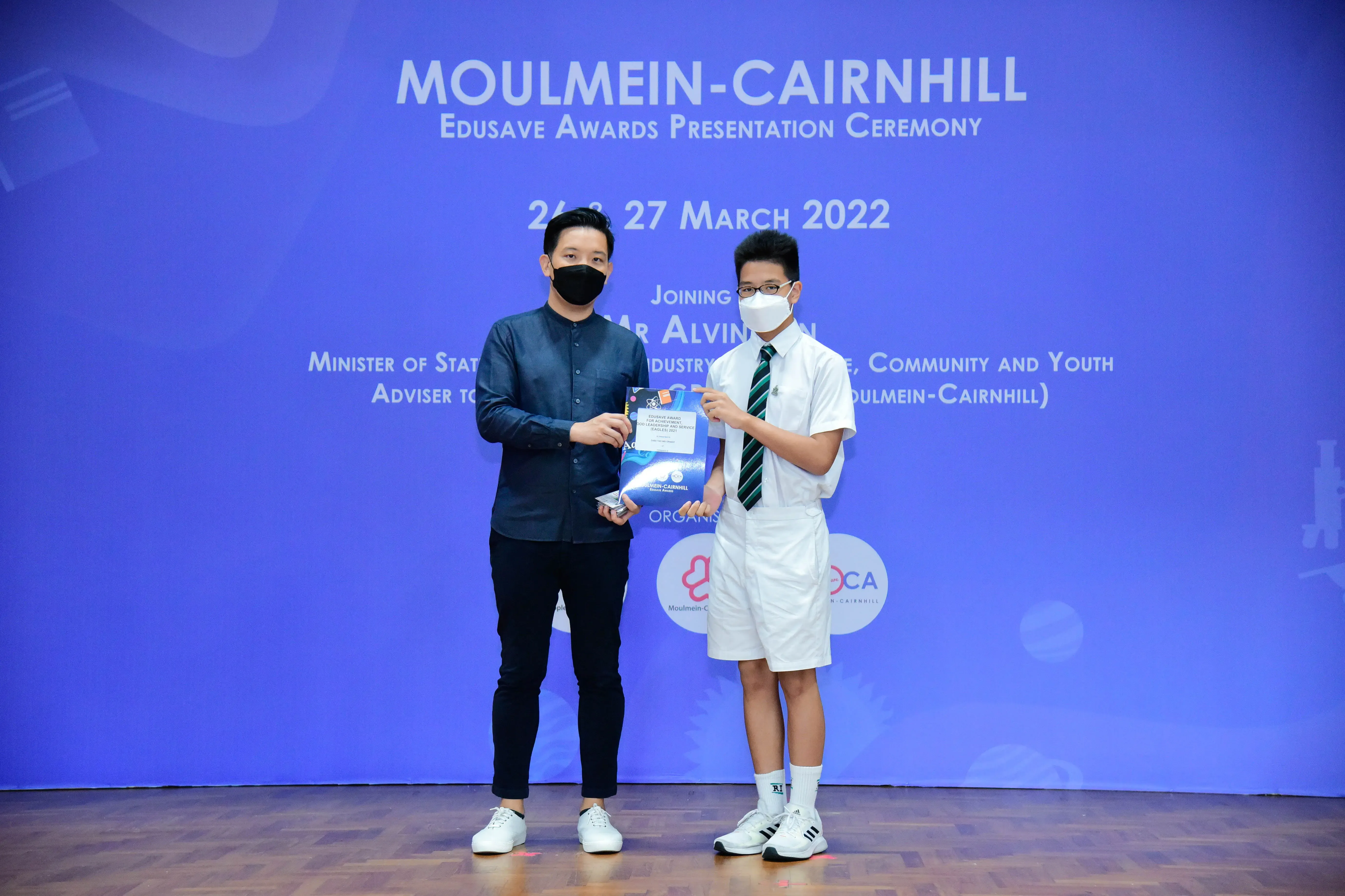 Two masked individuals hold an award at the Moulmein-Cairnhill Edusave Awards Presentation Ceremony, March 2022.