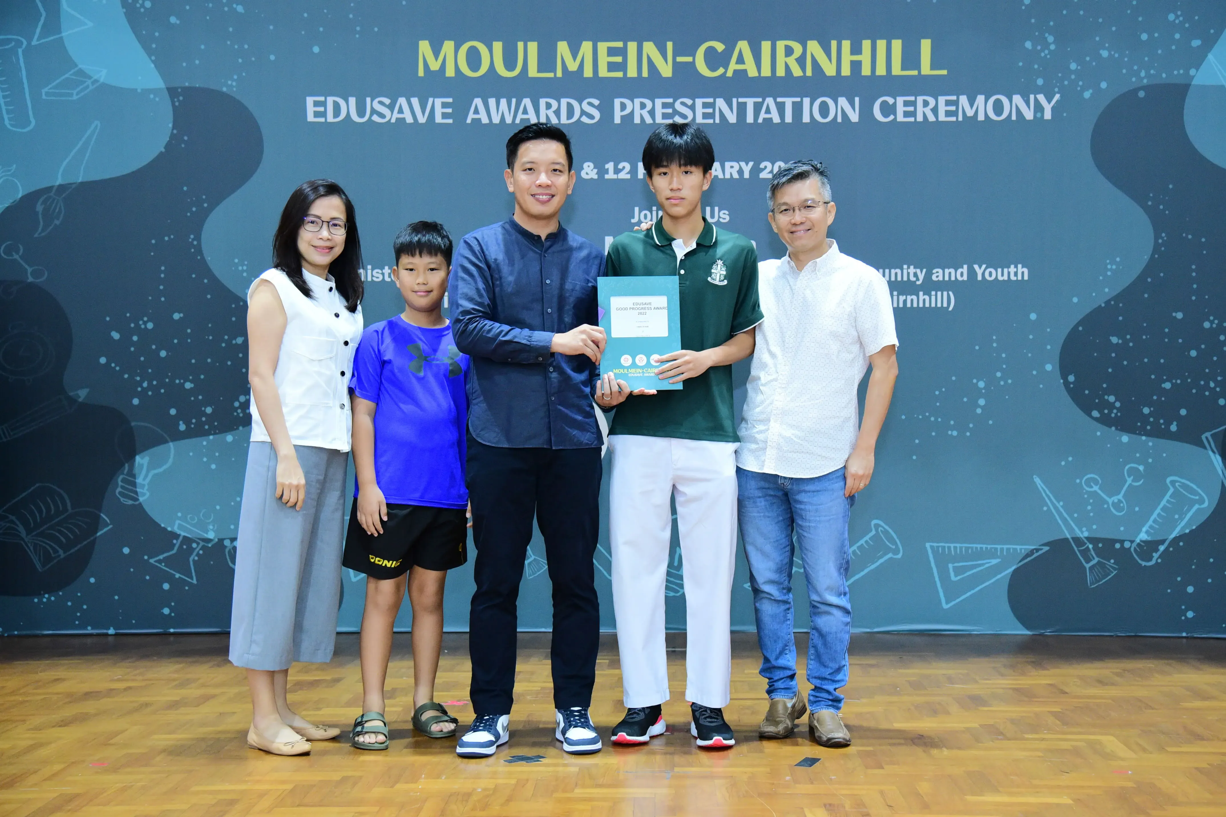 Five people pose on stage at the Moulmein-Cairnhill Edusave Awards Presentation Ceremony, with a teen holding a certificate.