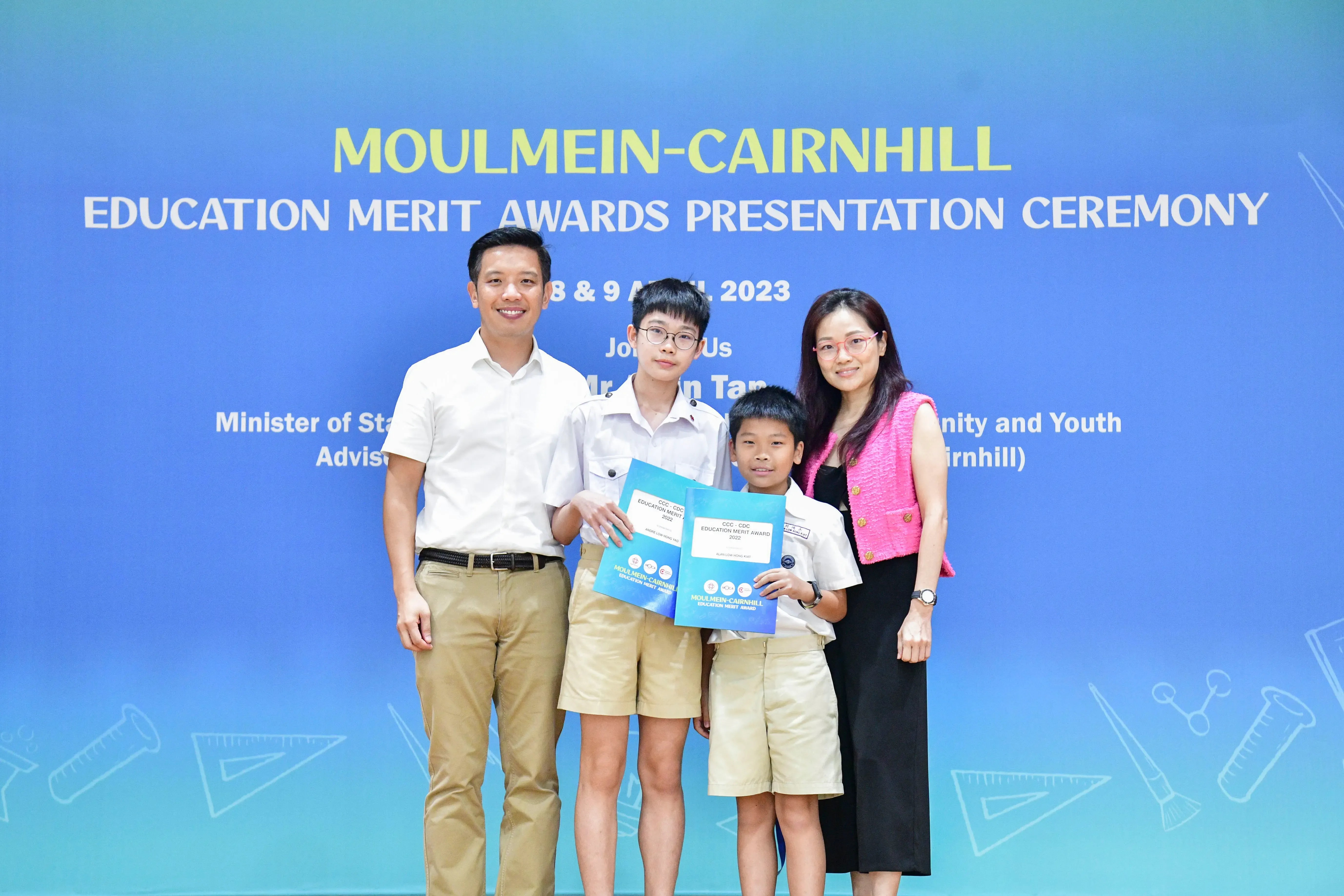 Two adults and two boys holding award folders pose before a Moulmein-Cairnhill Education Merit Awards banner.