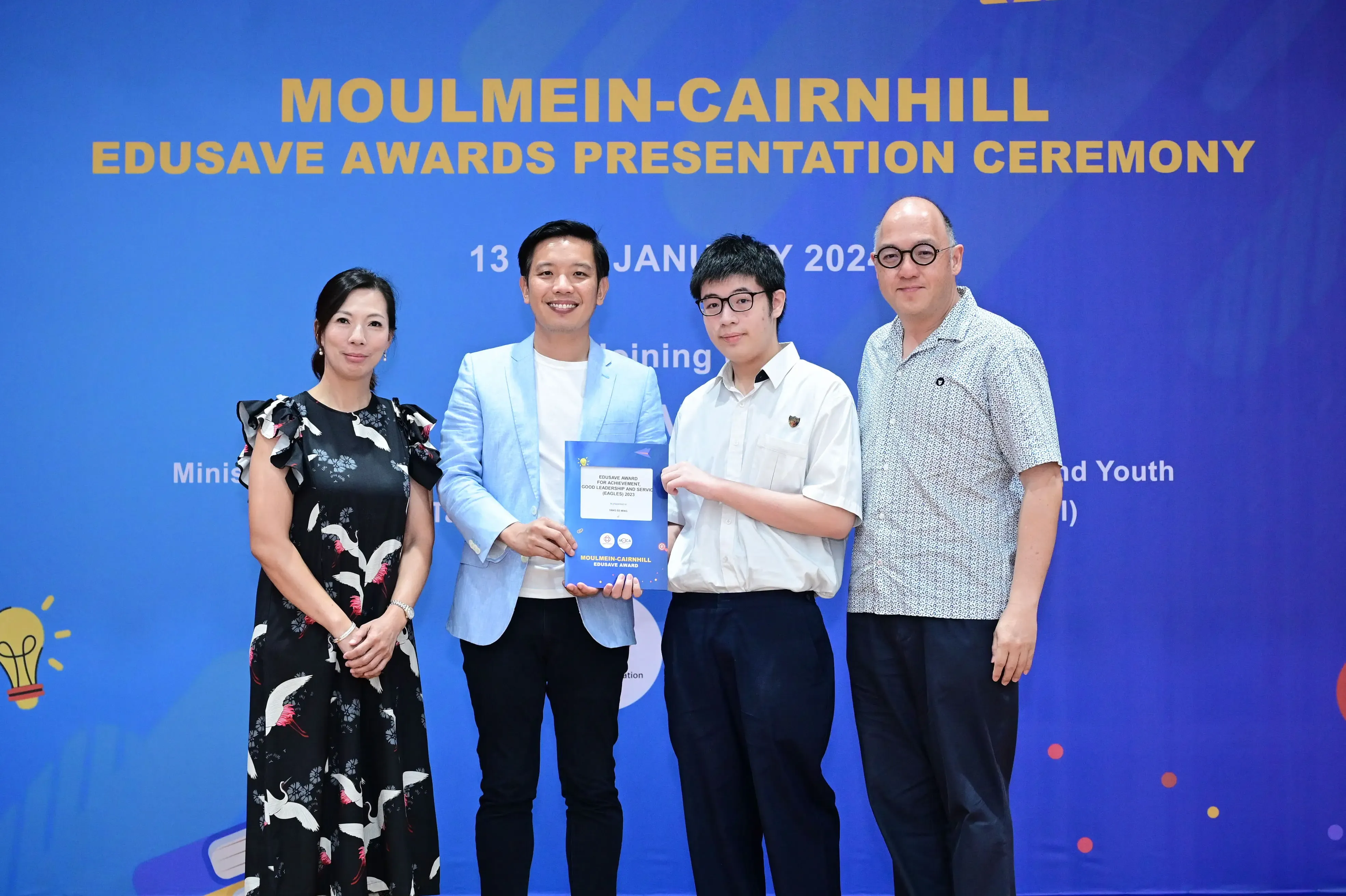 Four people pose at the Moulmein-Cairnhill Edusave Awards Presentation Ceremony, with a student holding a blue award certificate.