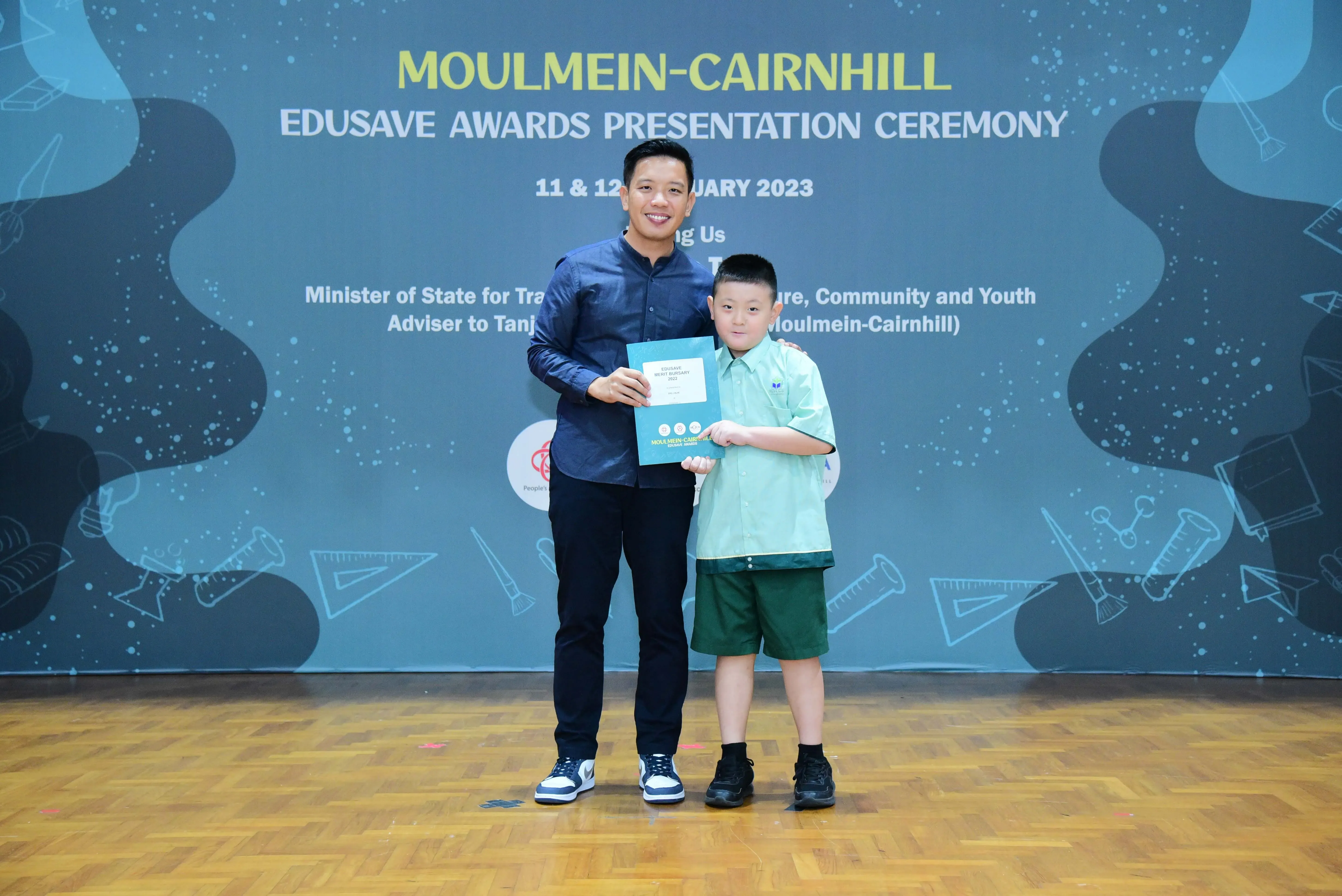 Adult and young boy hold a certificate at Moulmein-Cairnhill Edusave Awards Presentation Ceremony, January 2023.