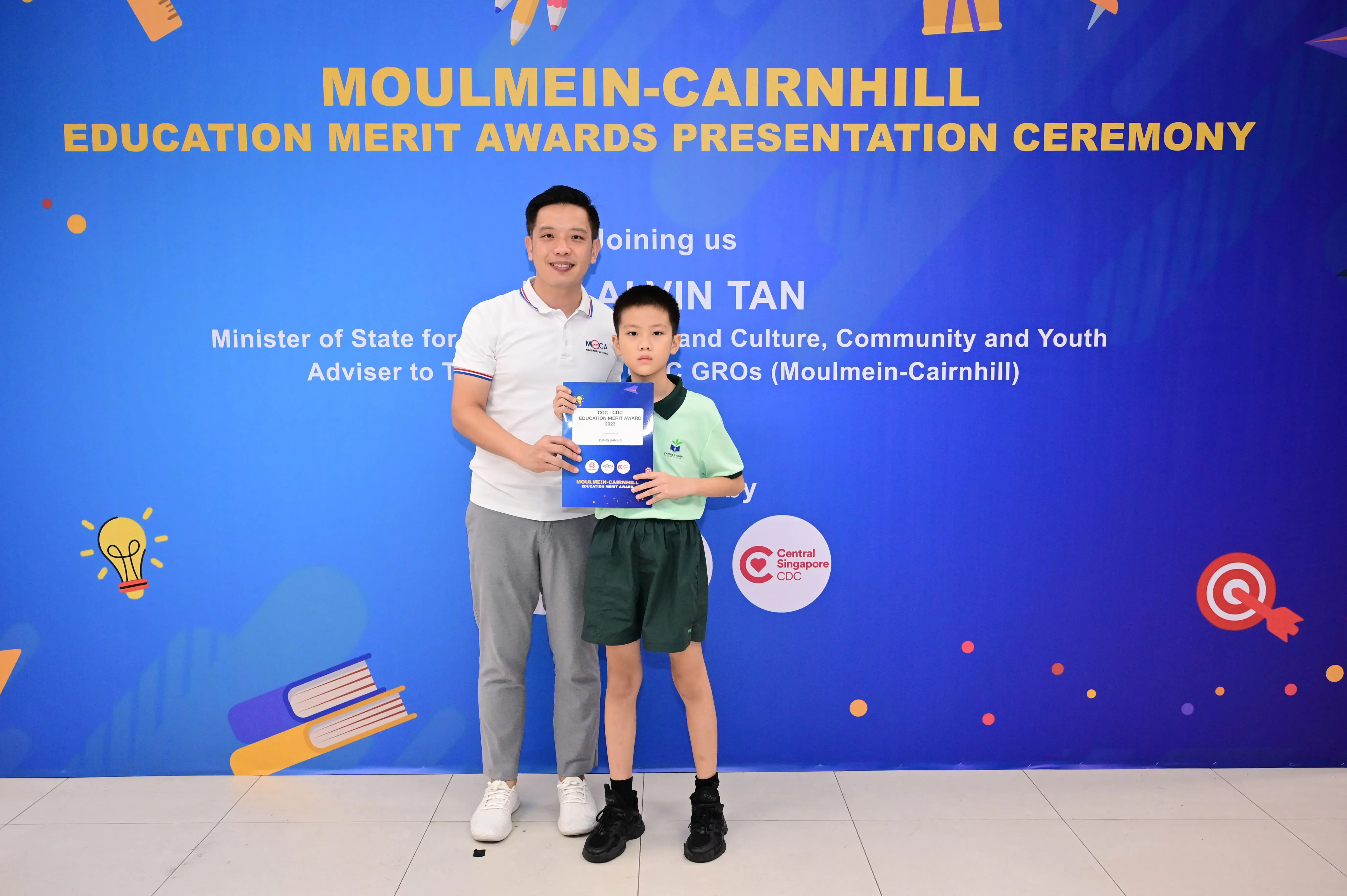 Man and young boy hold an award certificate in front of a Moulmein-Cairnhill Education Merit Awards backdrop.