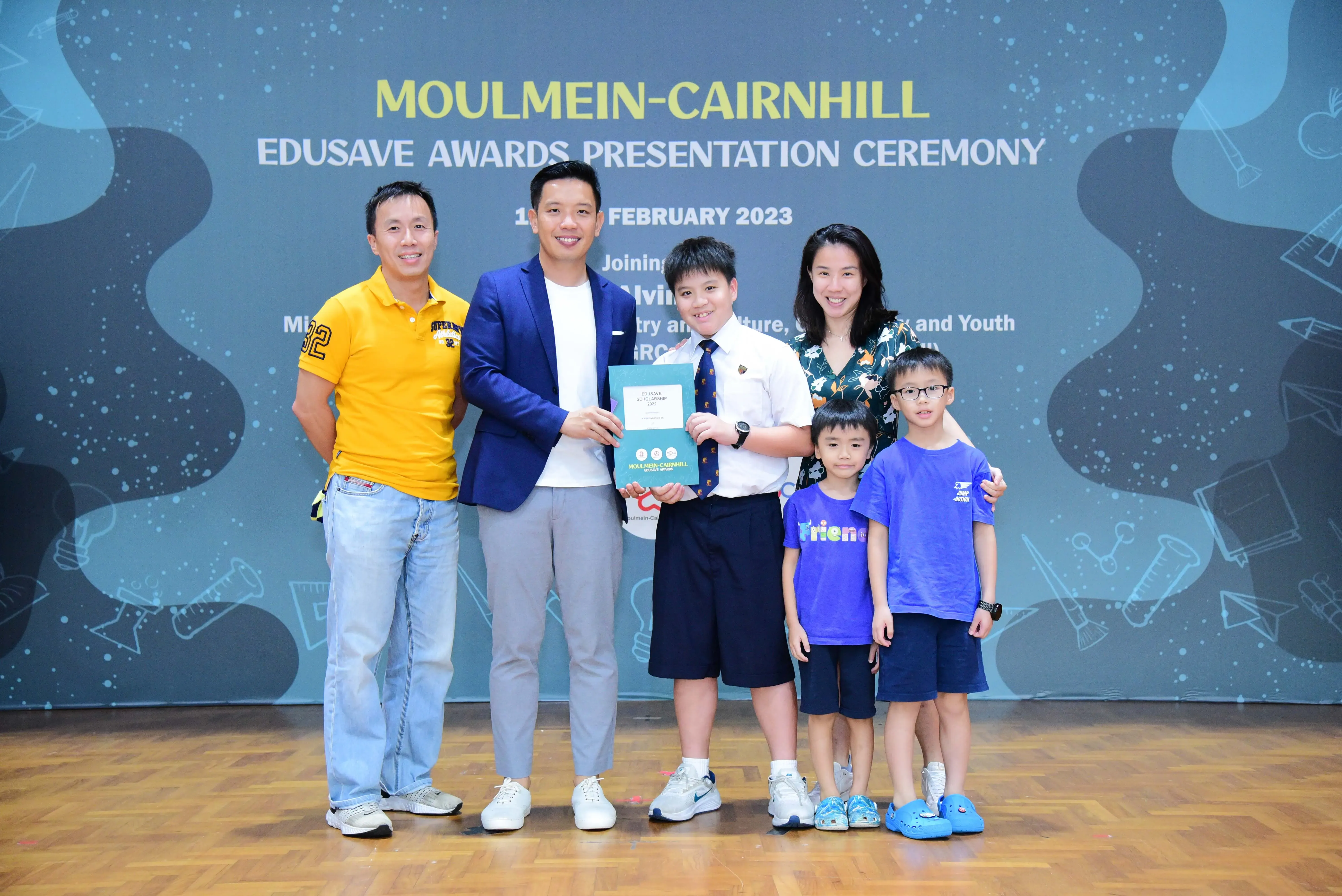 A boy holds an award certificate with adults and children at the Moulmein-Cairnhill Edusave Awards Ceremony, February 2023.