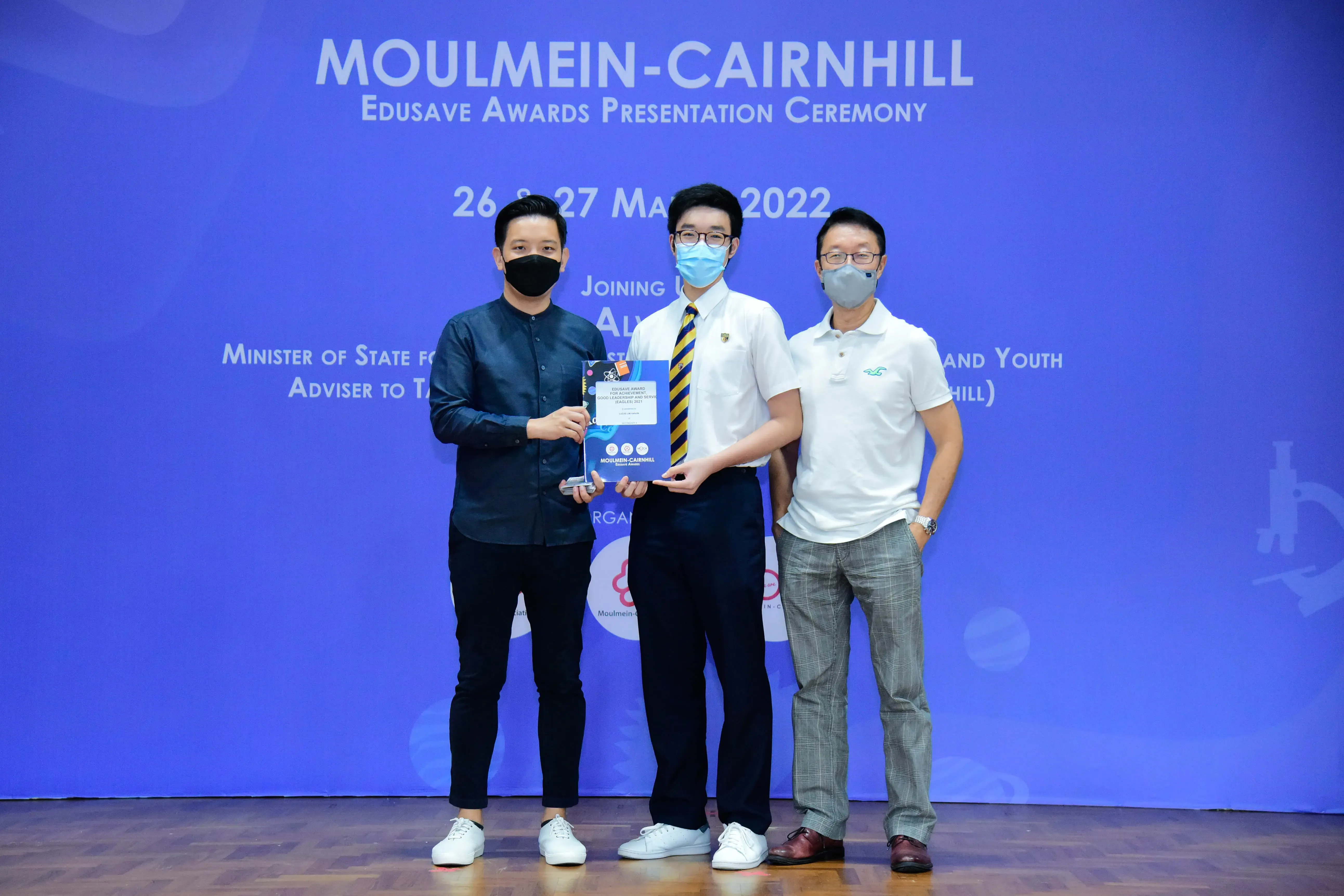 Three masked men pose at the Moulmein-Cairnhill Edusave Awards Presentation Ceremony in May 2022, holding an award.