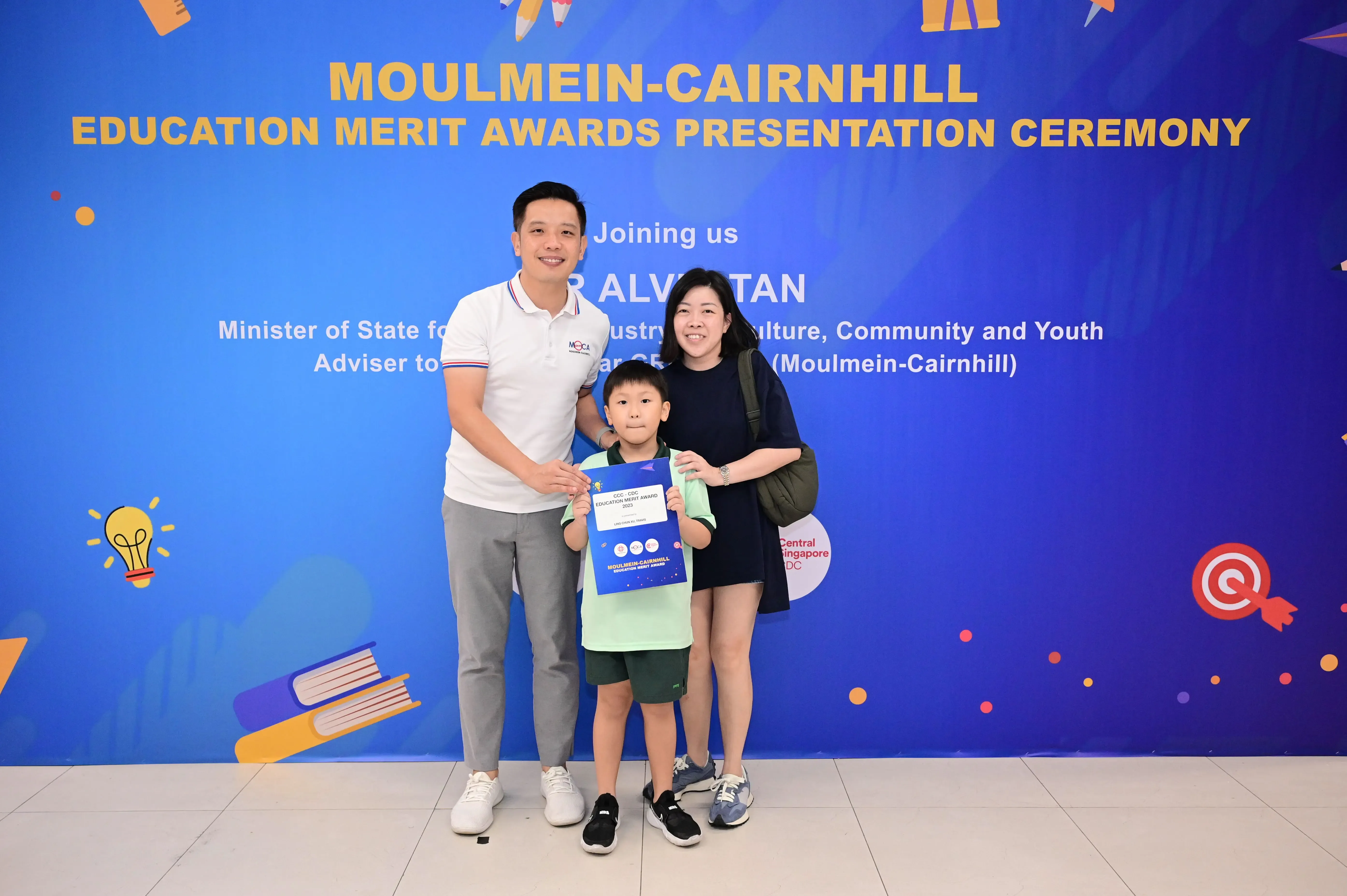 Two adults and a boy holding a certificate pose before a Moulmein-Cairnhill Education Merit Awards banner.