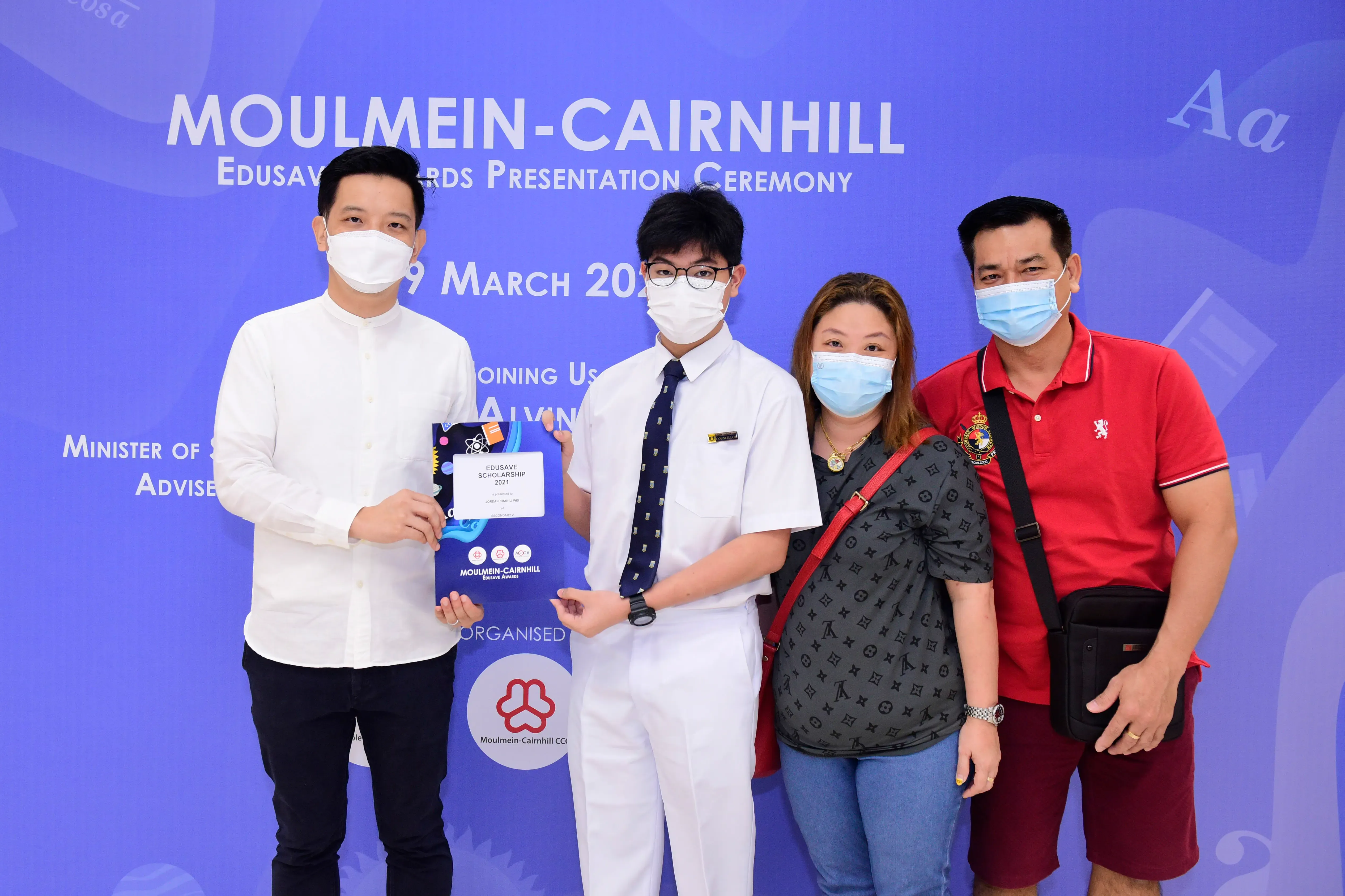 Four masked people pose at Moulmein-Cairnhill Edusave Awards Presentation Ceremony, with a student holding a certificate.