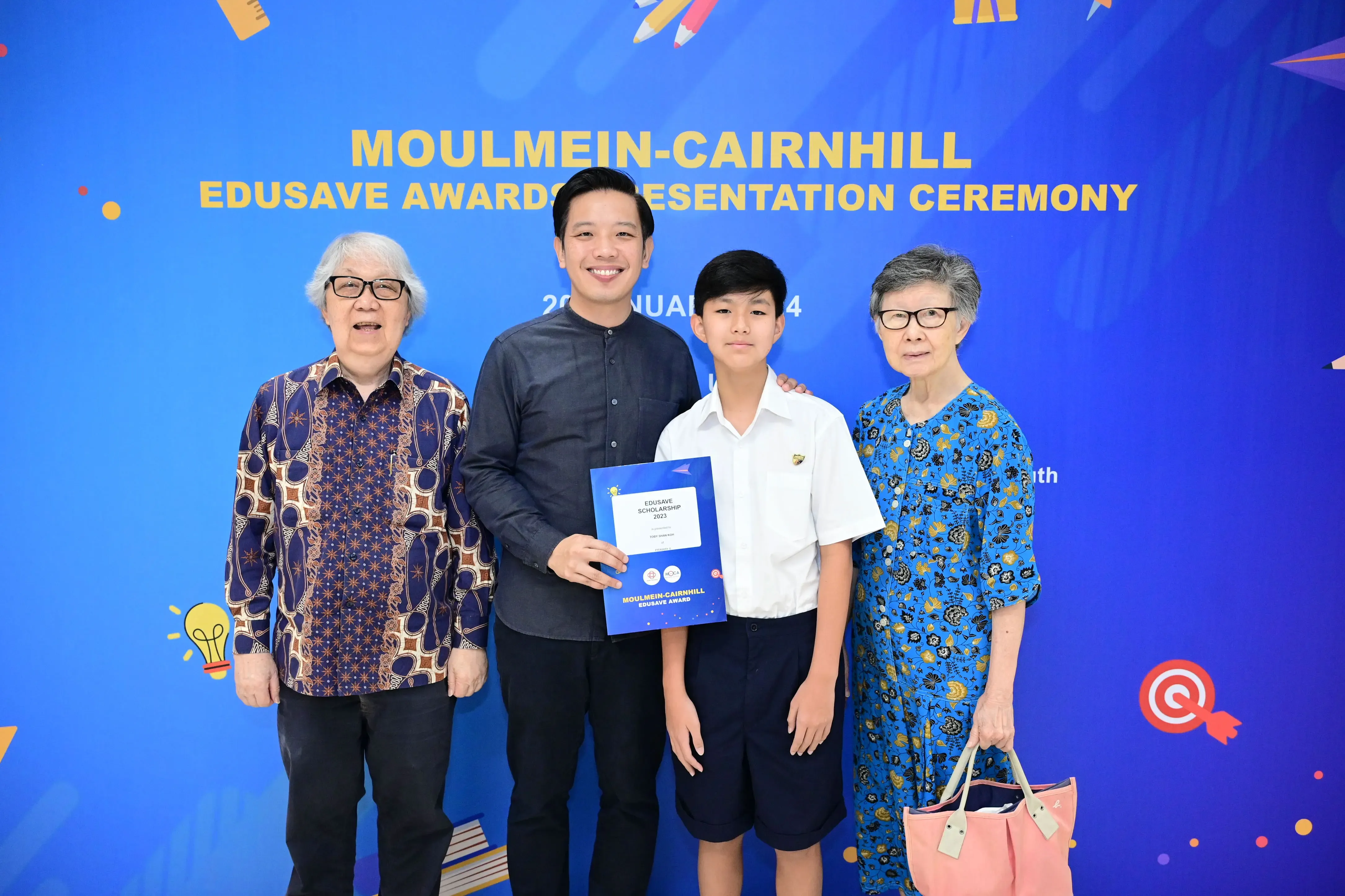 Four people pose in front of a Moulmein-Cairnhill Edusave Awards Presentation Ceremony banner, holding a certificate.