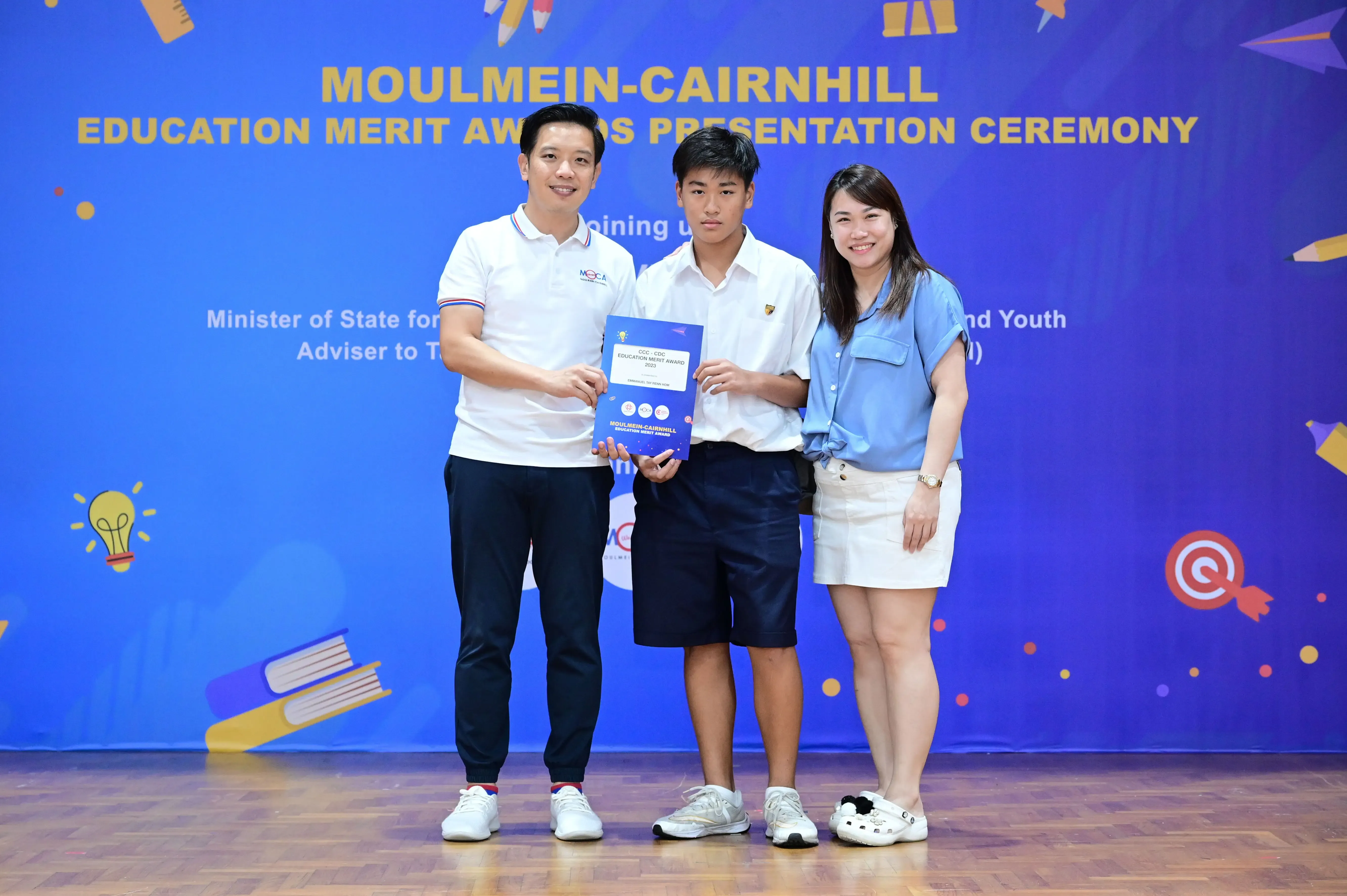 Three people pose holding a blue award booklet at the Moulmein-Cairnhill Education Merit Awards Presentation Ceremony.