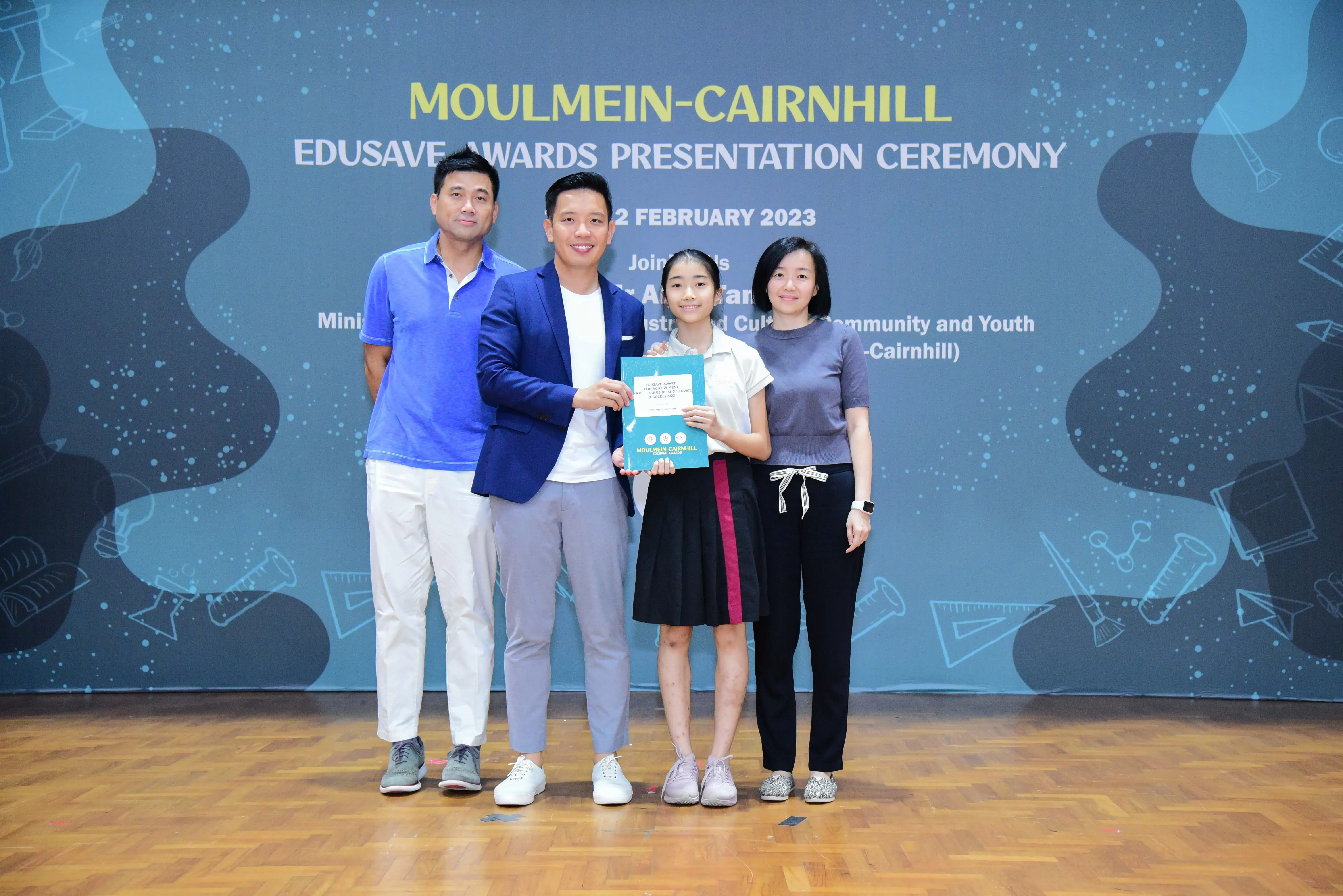 Four people pose together at the Moulmein-Cairnhill Edusave Awards Presentation Ceremony, with a girl holding a certificate.
