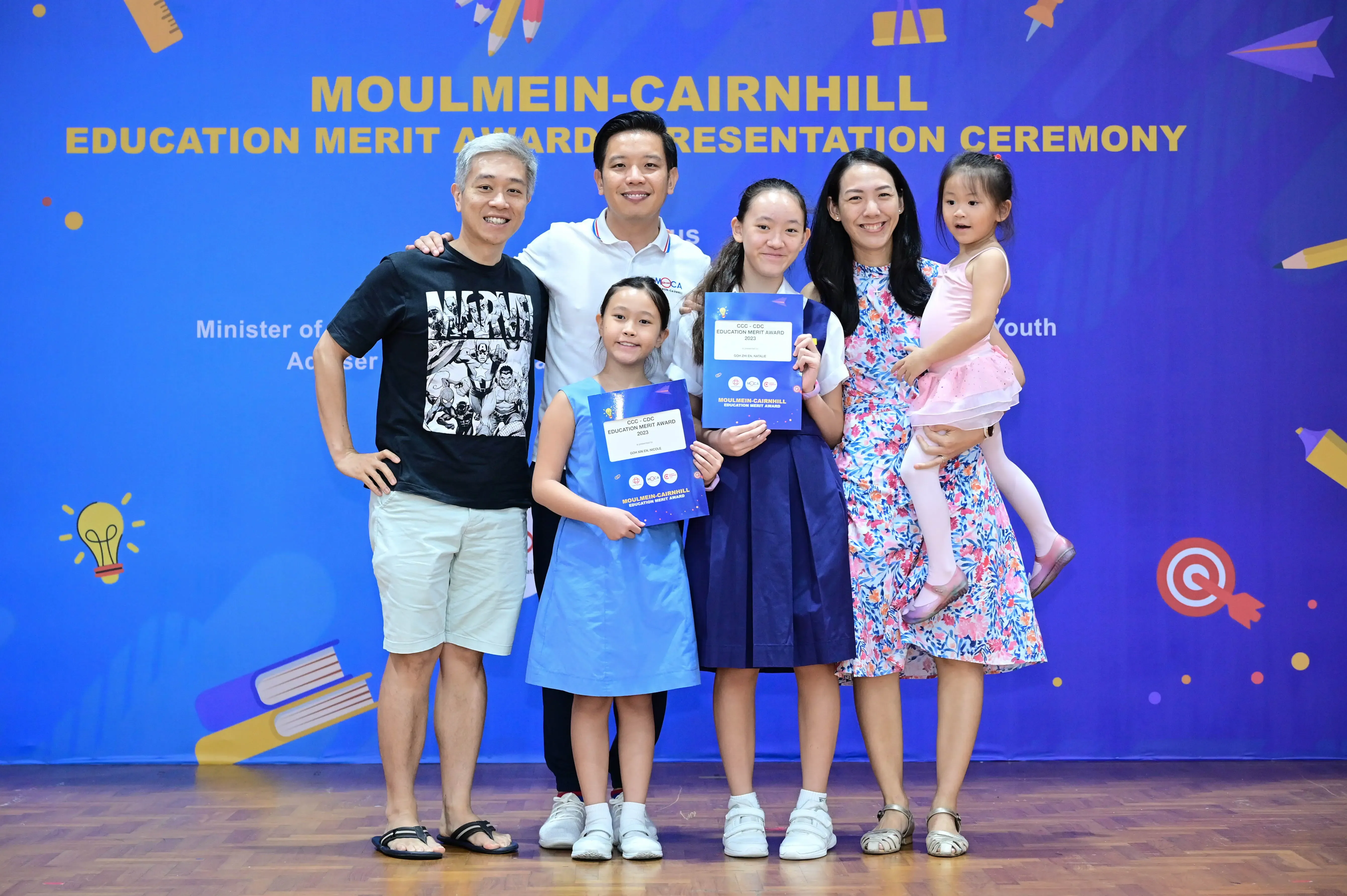A family of six poses at the Moulmein-Cairnhill Education Merit Award Presentation Ceremony, two girls holding award certificates.