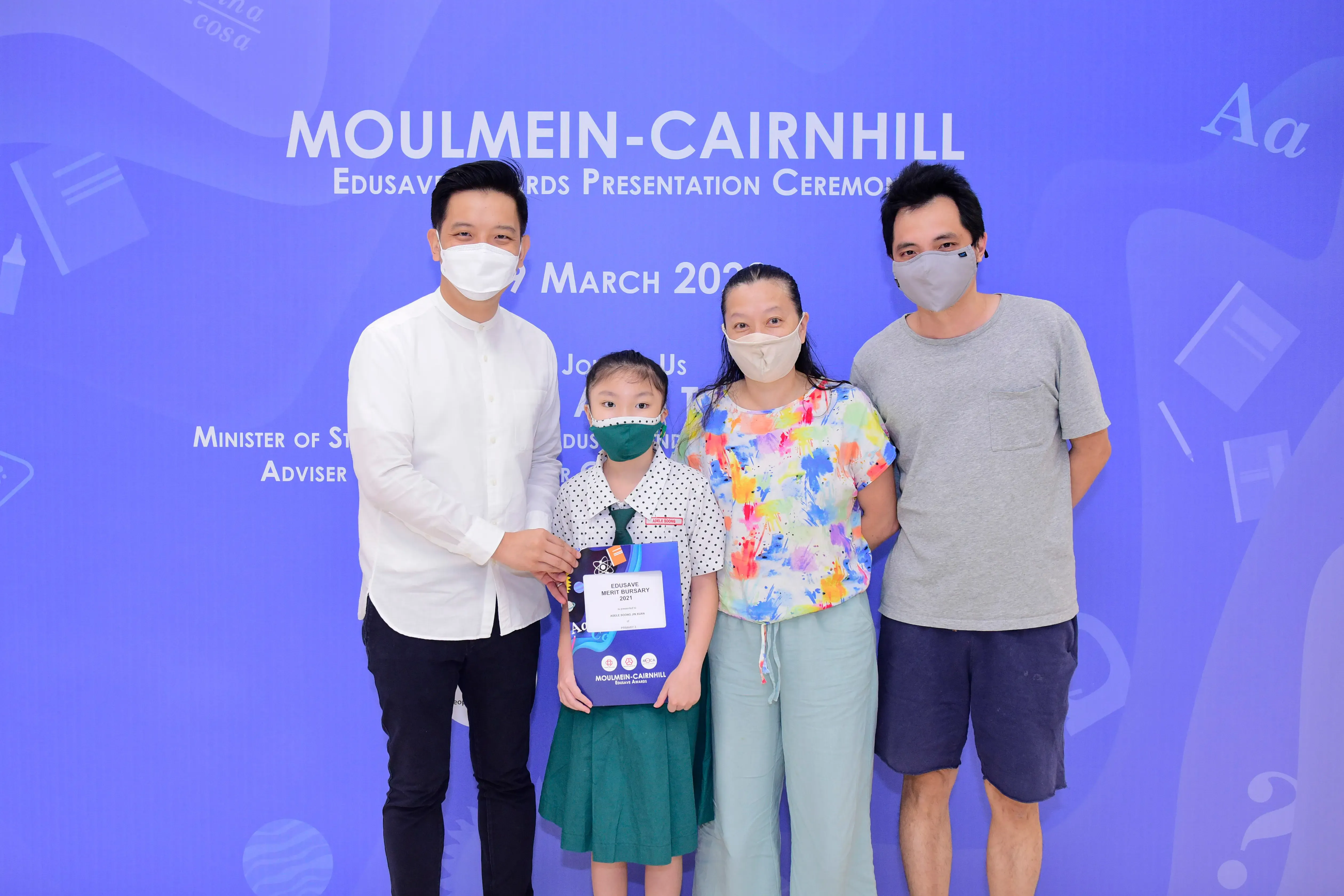 Four masked people pose before a Moulmein-Cairnhill Edusave Awards ceremony backdrop, with a girl holding a bursary certificate.