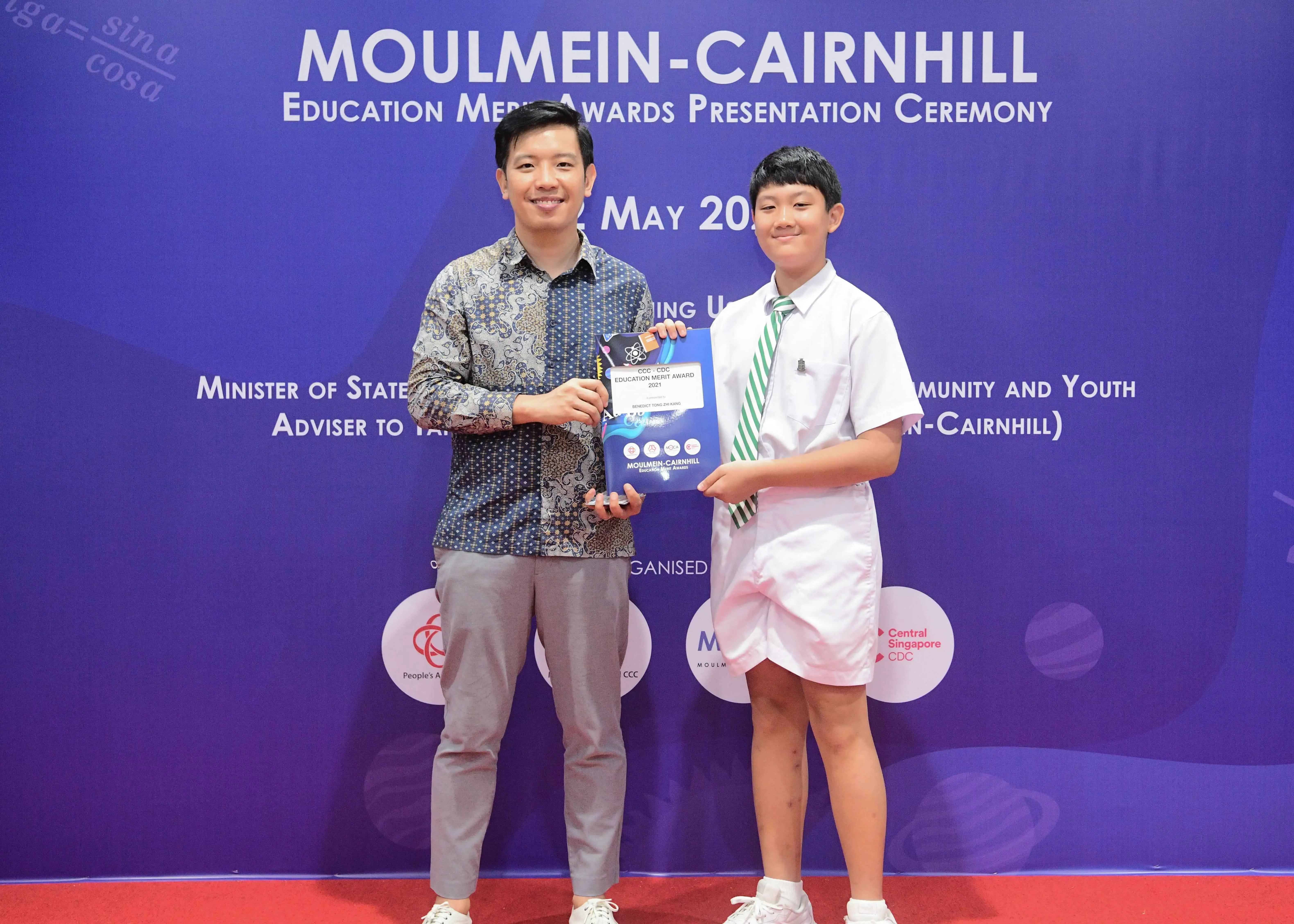 An adult and a young student hold an award together at the Moulmein-Cairnhill Education Merit Awards ceremony.