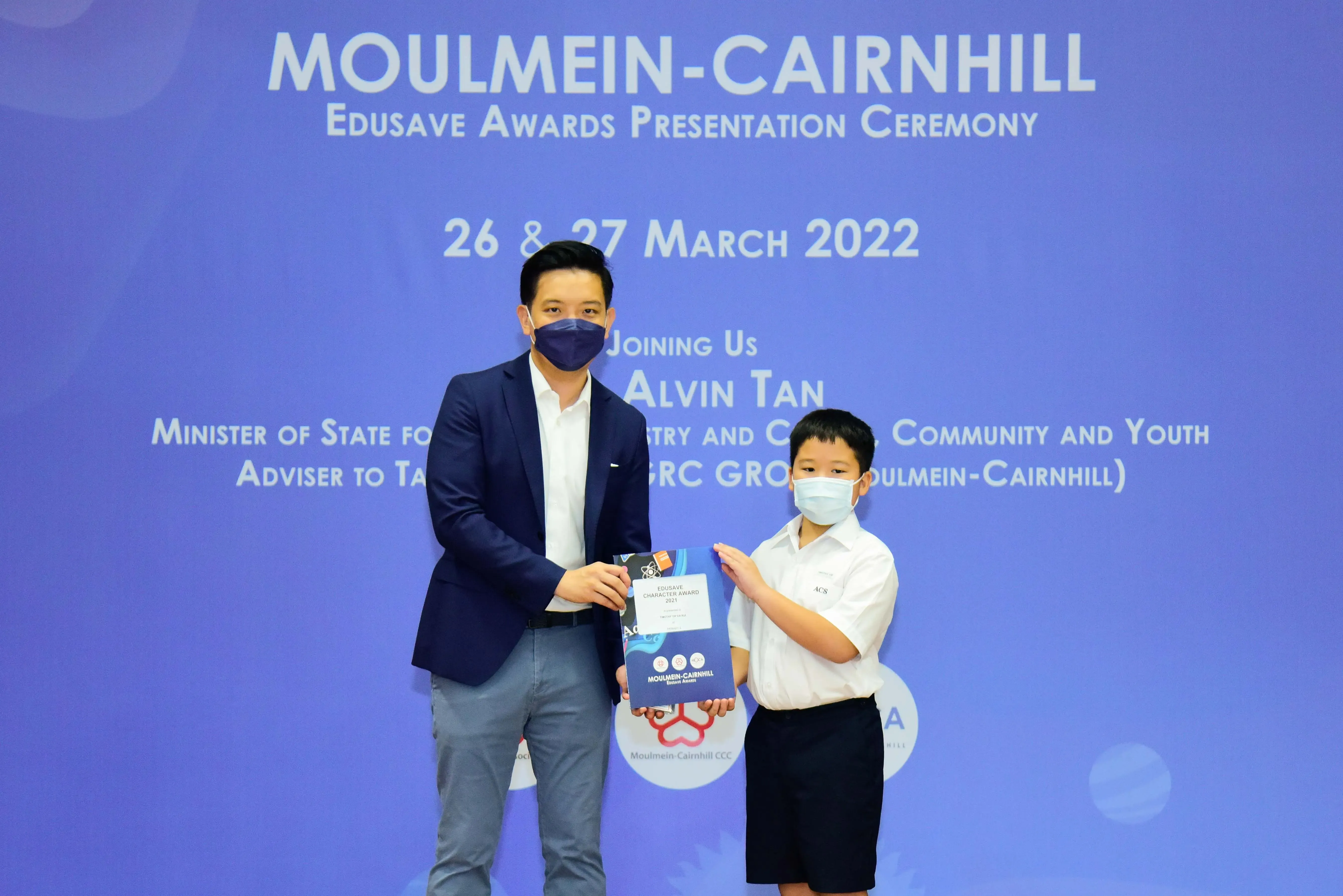 An adult and a young boy, both masked, hold an award at the Moulmein-Cairnhill Edusave Awards Ceremony, March 2022.