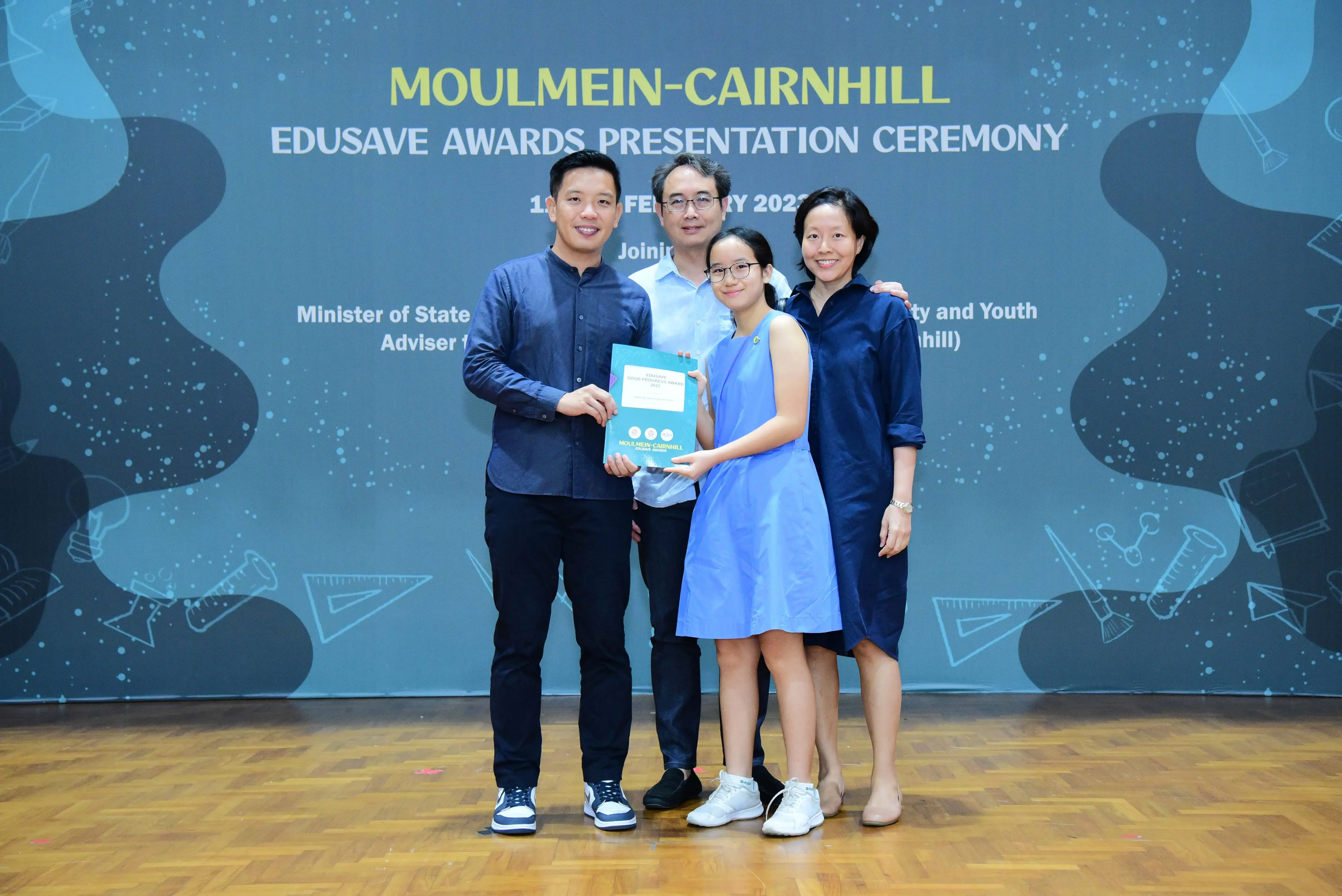 Four people pose together at the Moulmein-Cairnhill Edusave Awards Presentation Ceremony, with a girl holding an award folder.