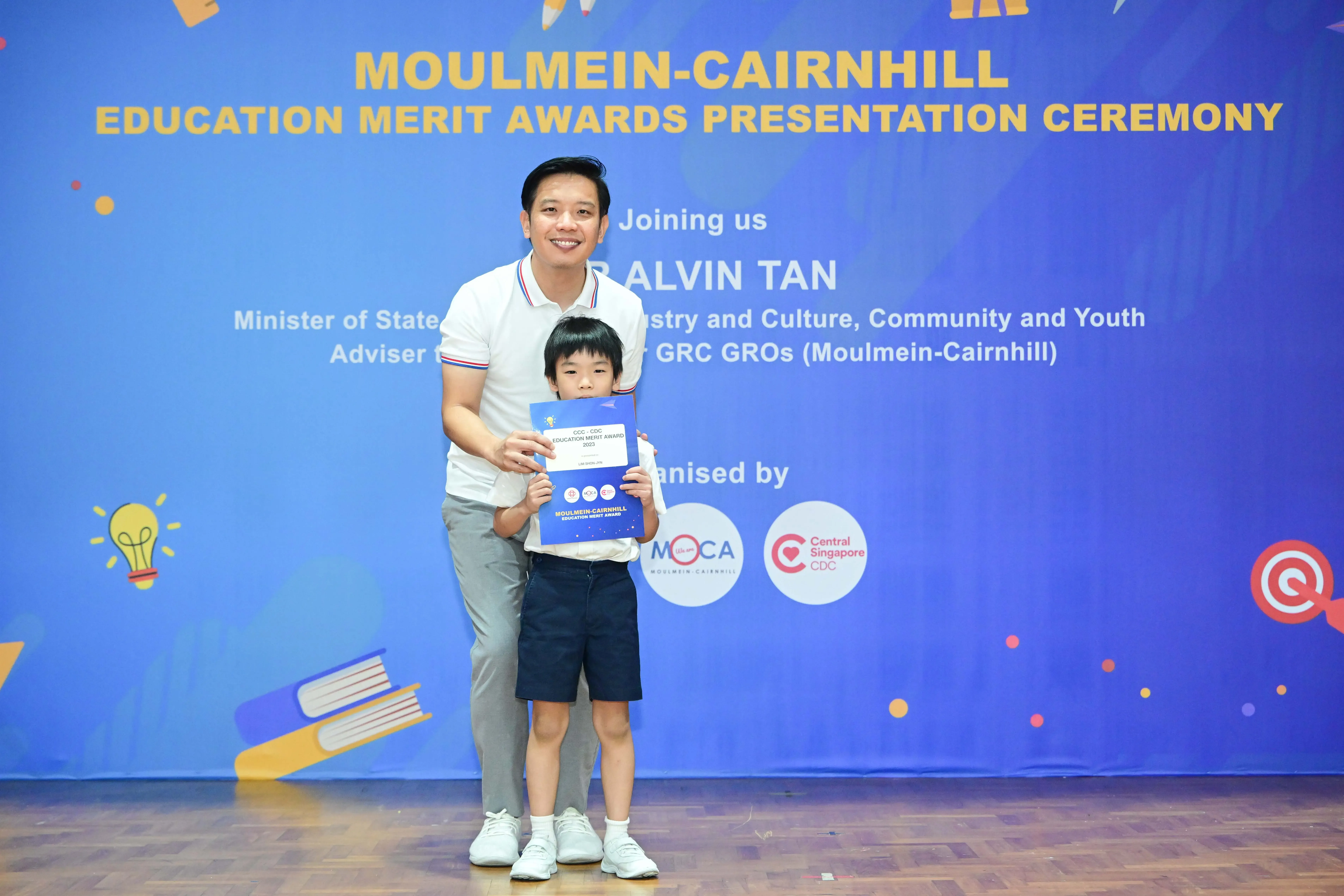 A man and young boy holding a certificate stand before a Moulmein-Cairnhill Education Merit Awards banner.
