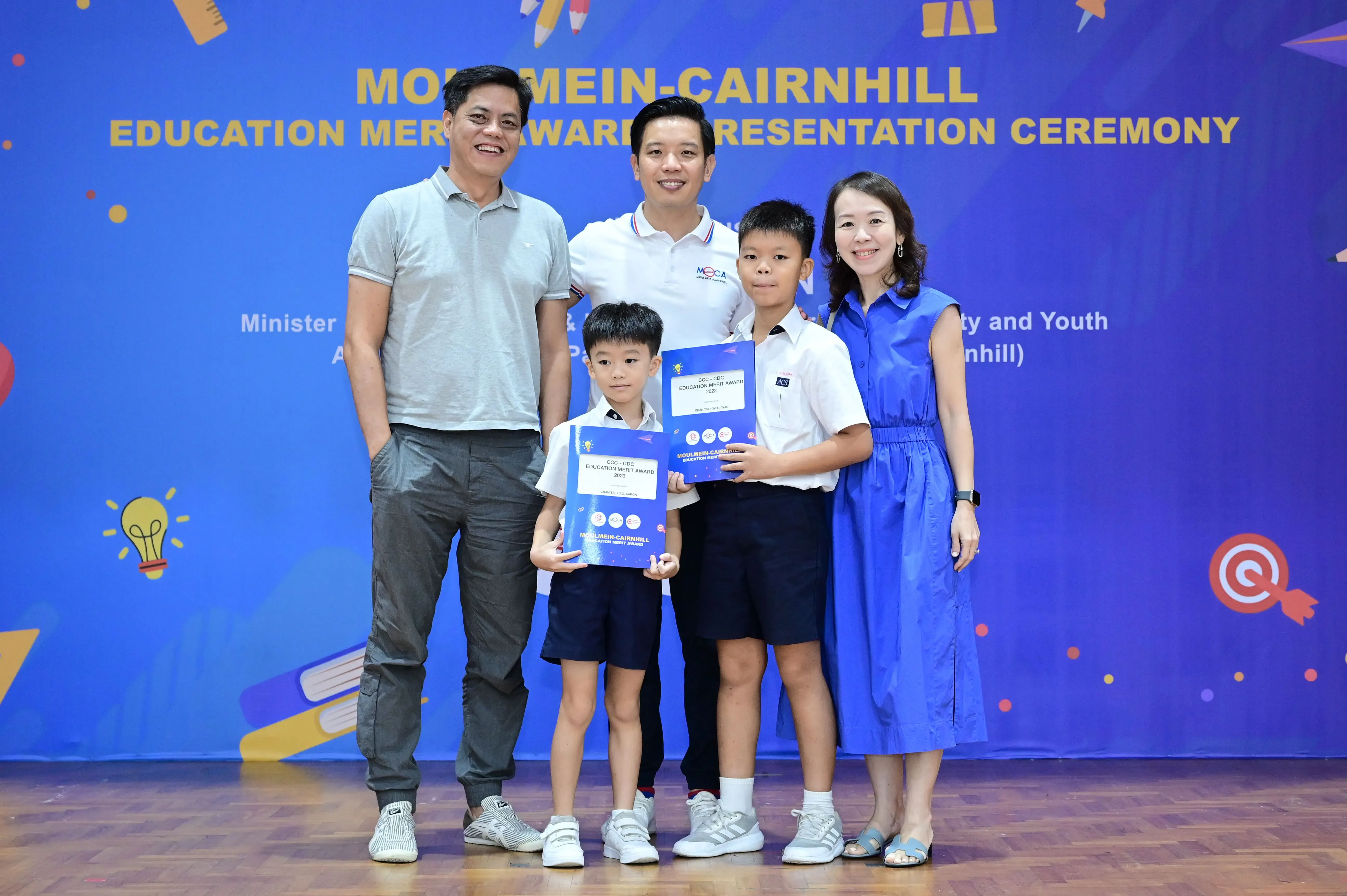 Two boys holding award folders pose with three adults at a Moulmein-Cairnhill Education Merit Award ceremony.