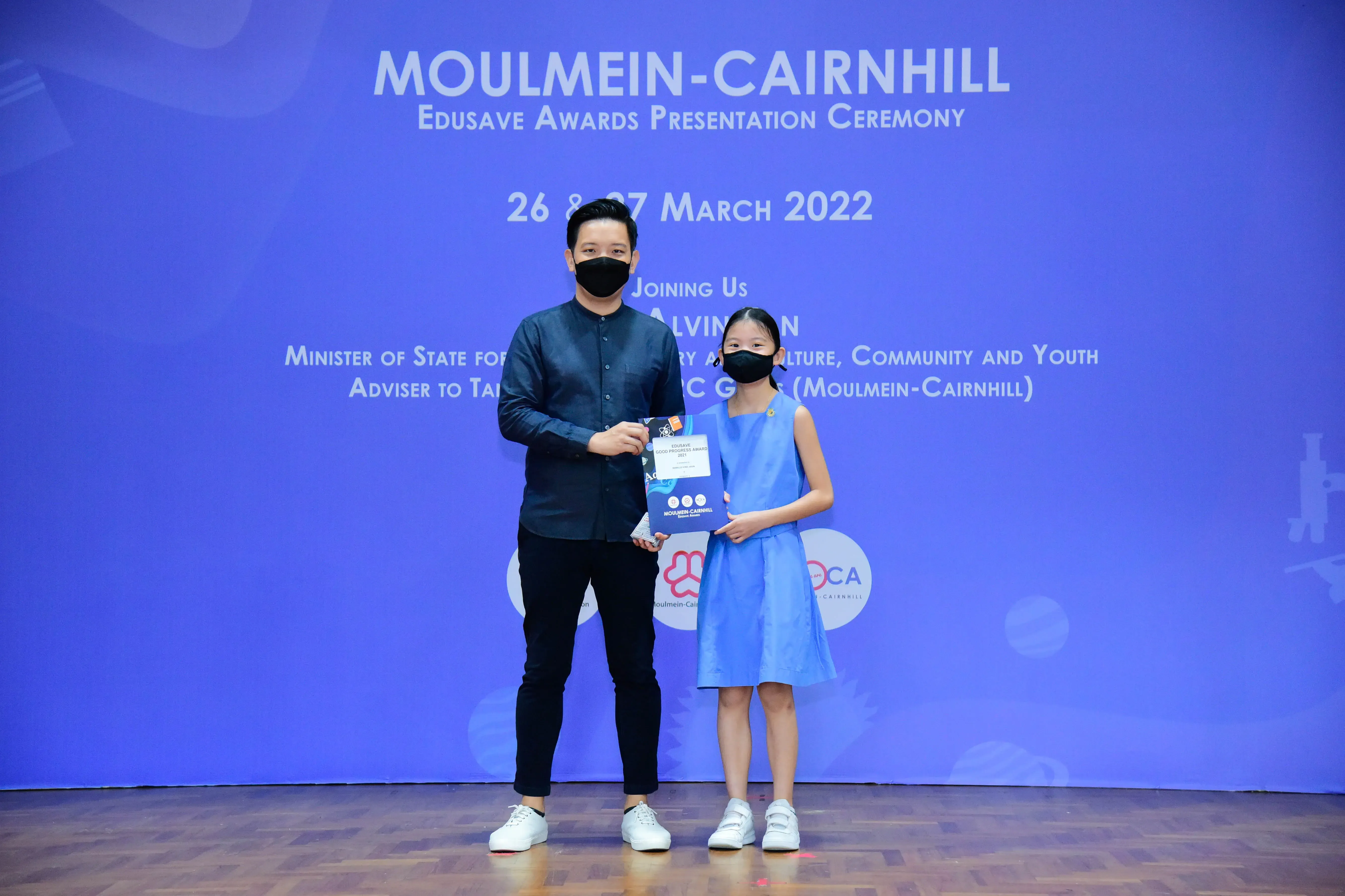 An adult and a young girl wearing masks hold an award certificate at the Moulmein-Cairnhill Edusave Awards Ceremony 2022.