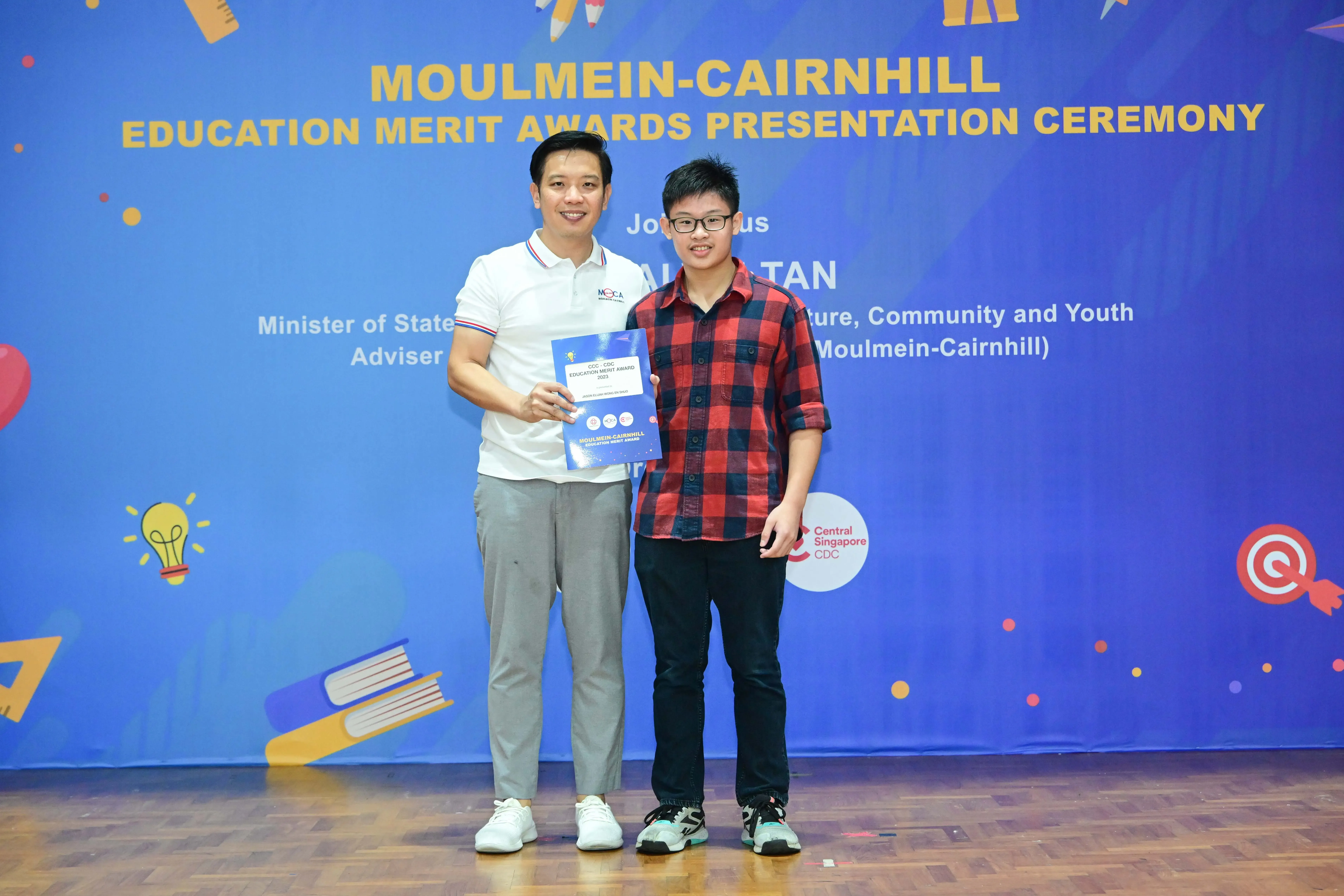 Two people stand before a blue Moulmein-Cairnhill Education Merit Awards banner, holding a certificate together.