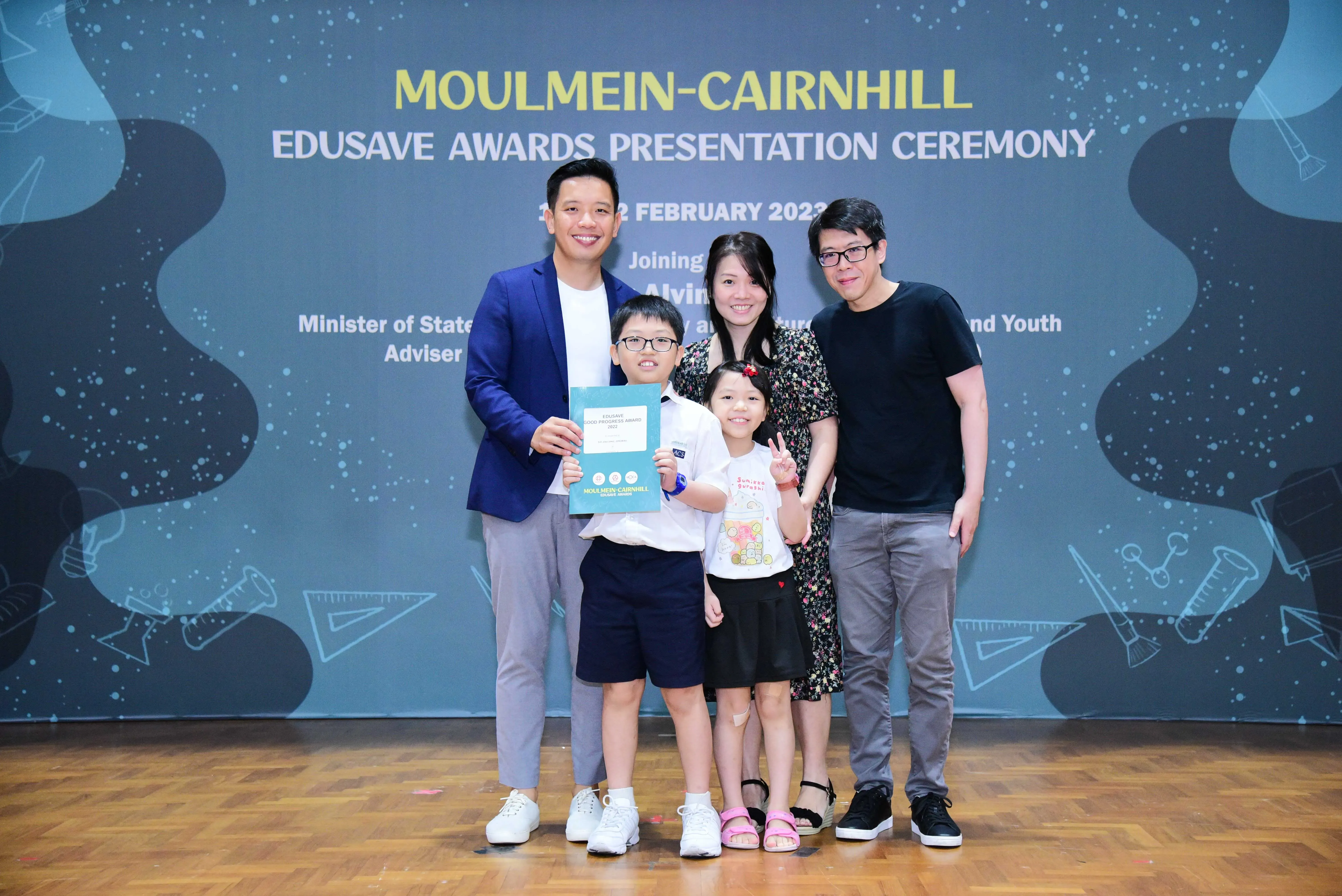 Five people pose at the Moulmein-Cairnhill Edusave Awards Presentation Ceremony; a boy holds a teal award certificate.