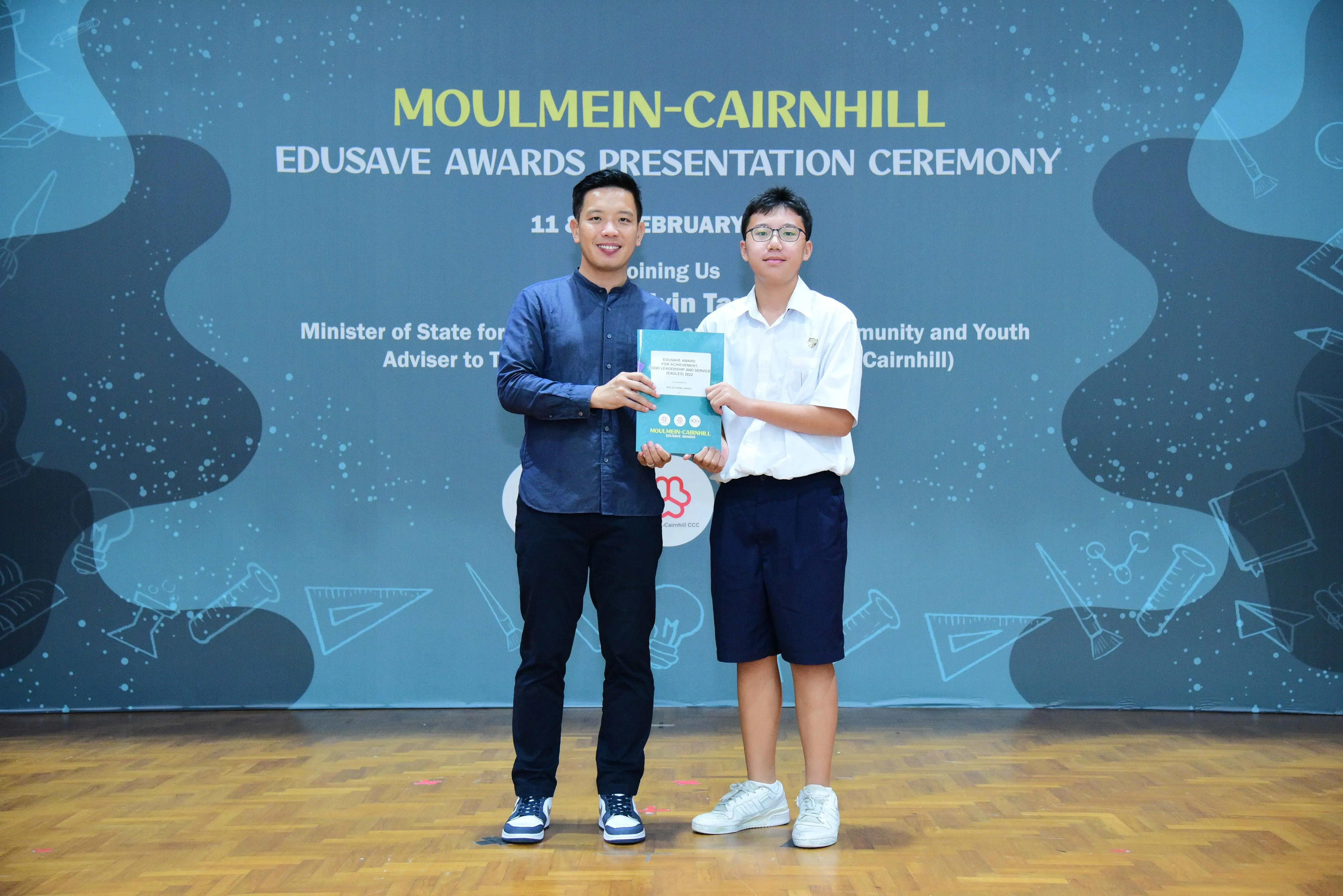 Two people hold a certificate together at the Moulmein-Cairnhill Edusave Awards Presentation Ceremony backdrop.