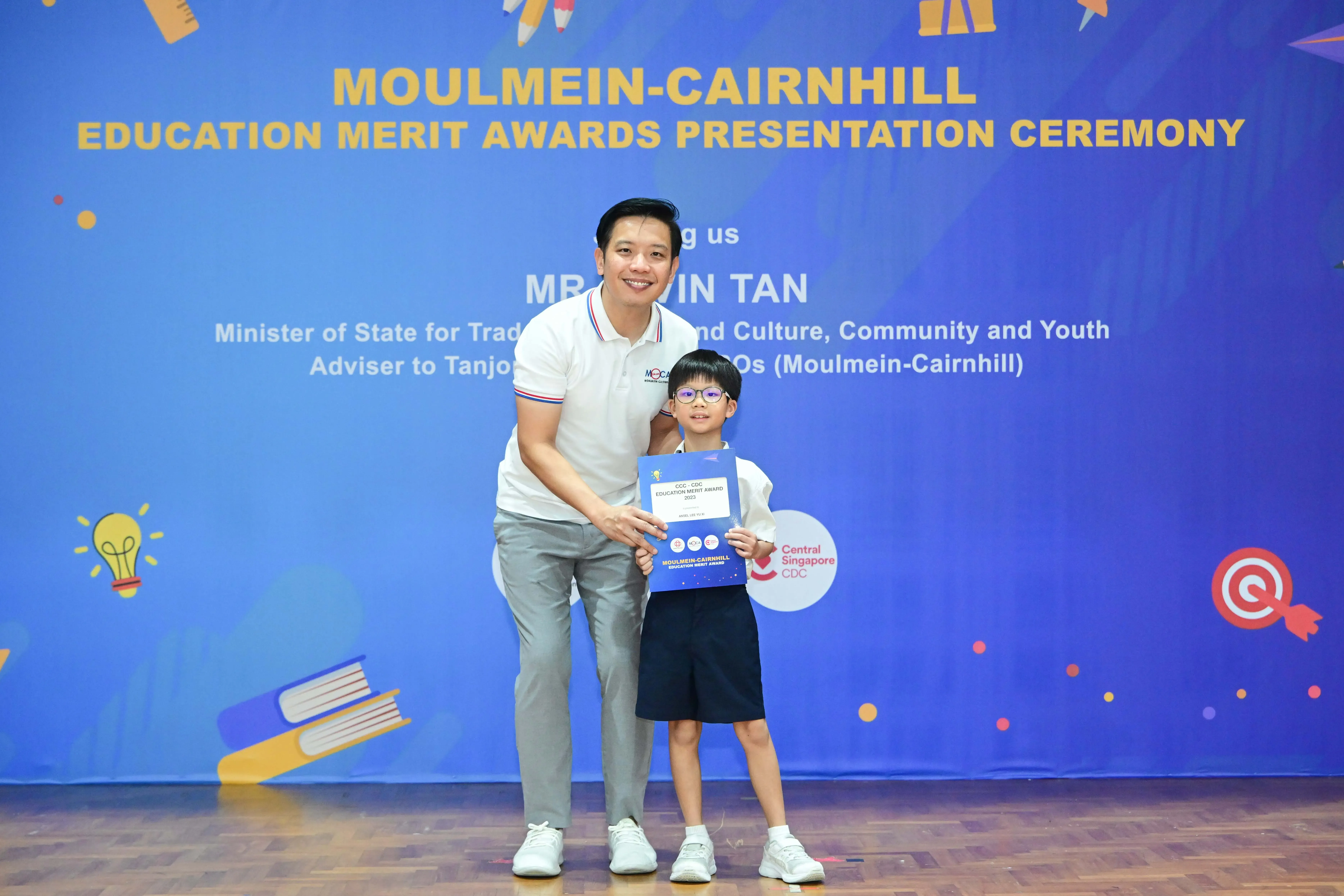 A man and a young boy holding a merit award certificate in front of a Moulmein-Cairnhill Education Merit Awards banner.