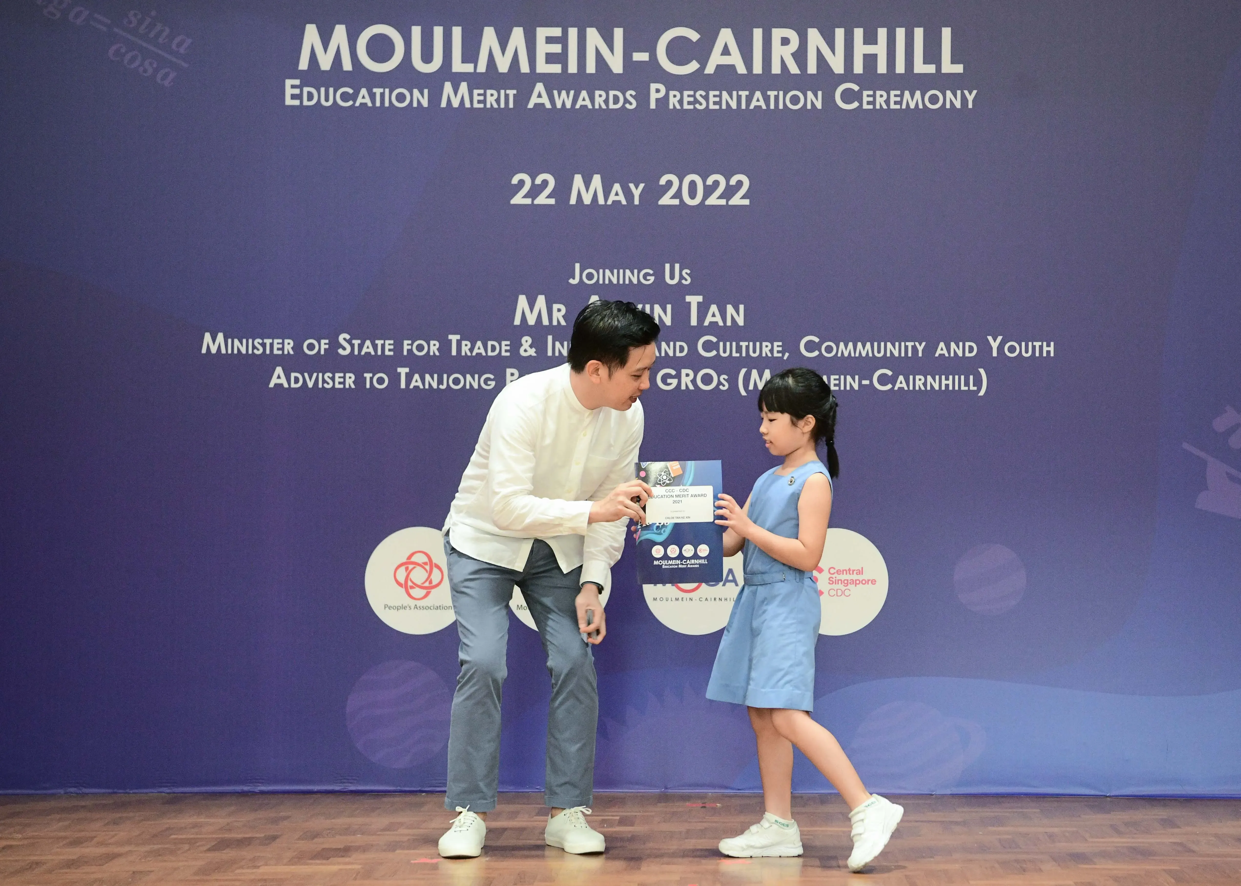 A man in white shirt hands a certificate to a young girl at the Moulmein-Cairnhill Education Merit Awards, 22 May 2022.