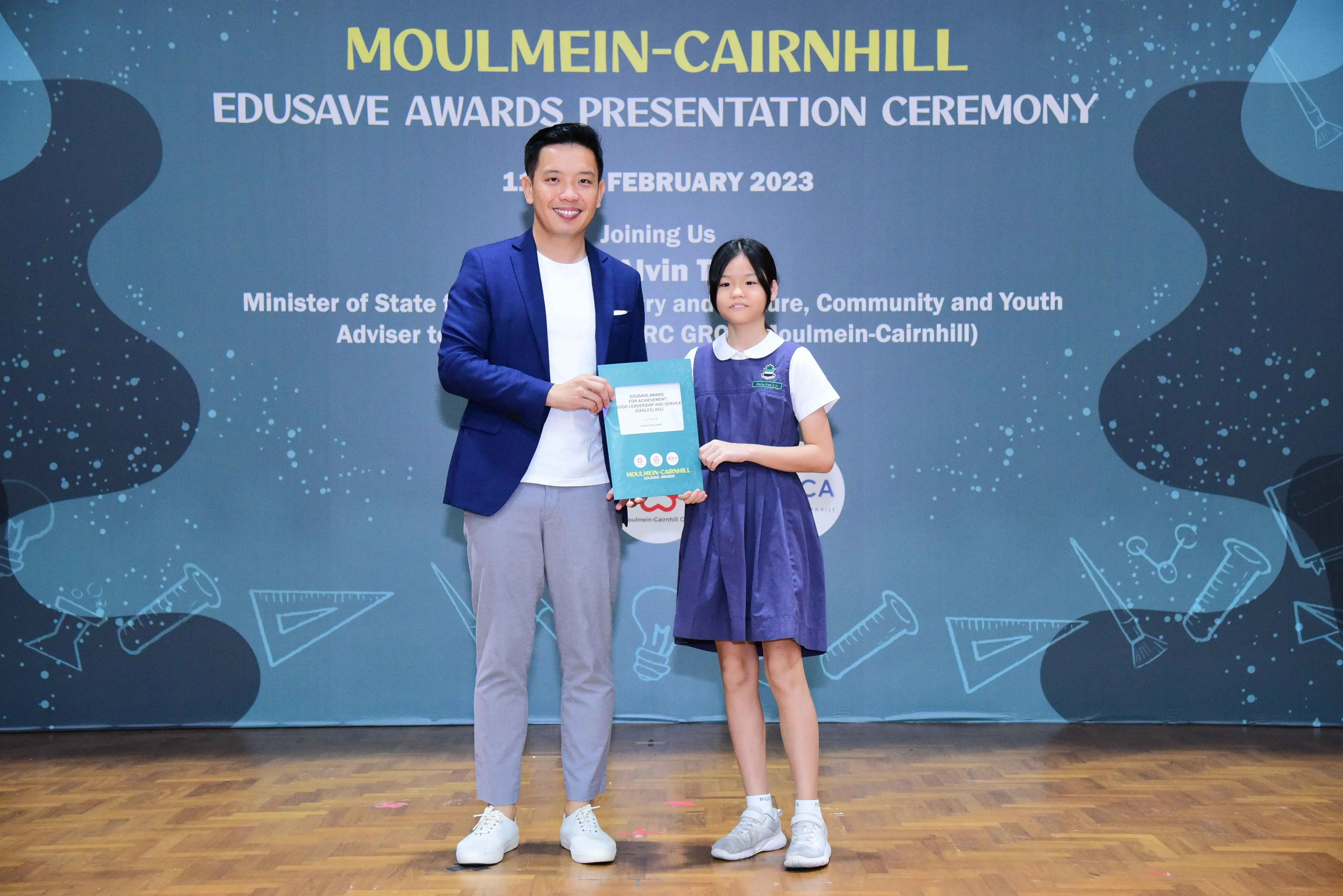 A man in a blue blazer and a young girl in school uniform hold an award at the Moulmein-Cairnhill Edusave Awards 2023.