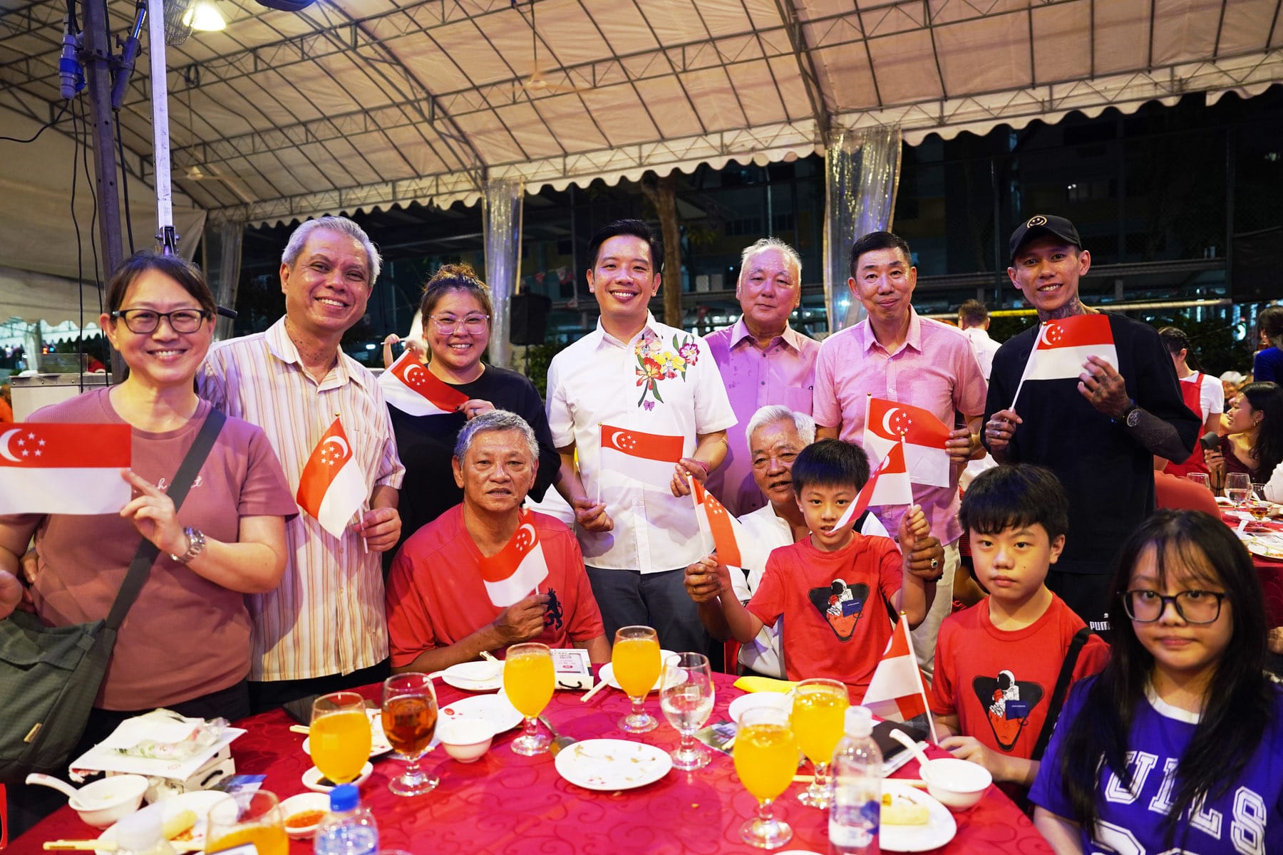 A group of smiling people holding small Singapore flags at an outdoor dinner event under a tent at night.