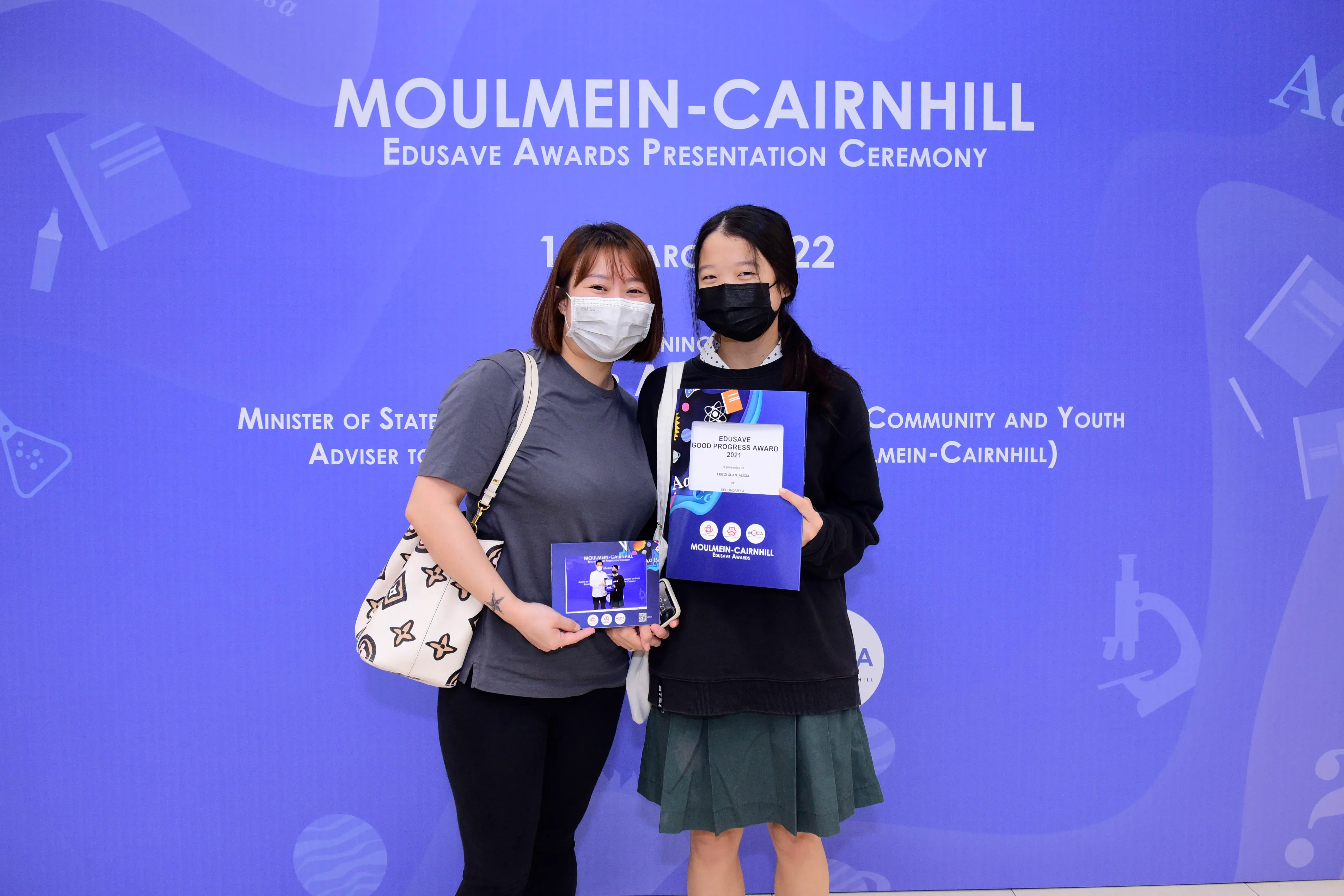 Two masked women pose holding an Edusave Good Progress Award certificate at the Moulmein-Cairnhill ceremony backdrop.