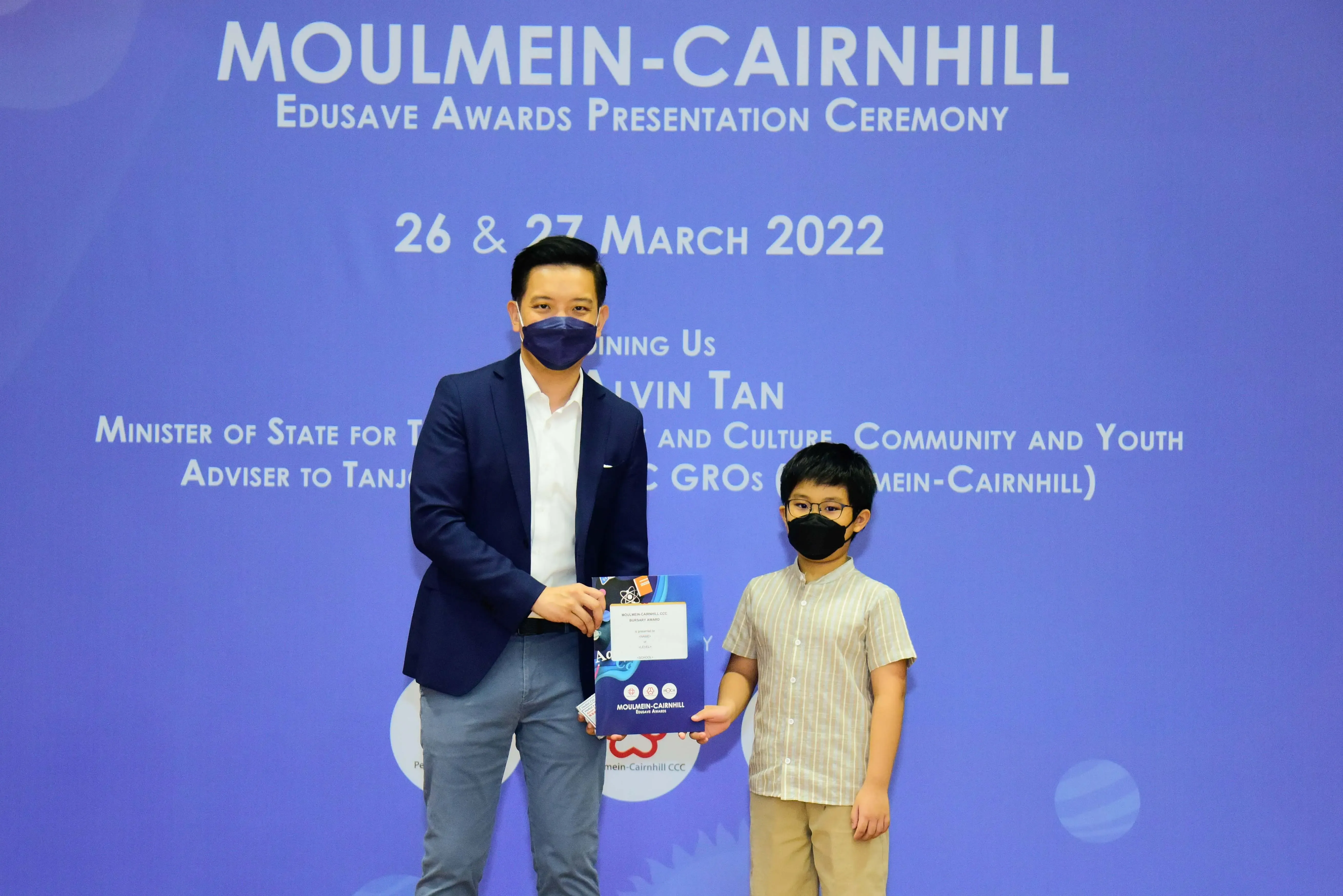 A masked adult and young boy hold an award folder at the Moulmein-Cairnhill Edusave Awards Ceremony, March 2022.