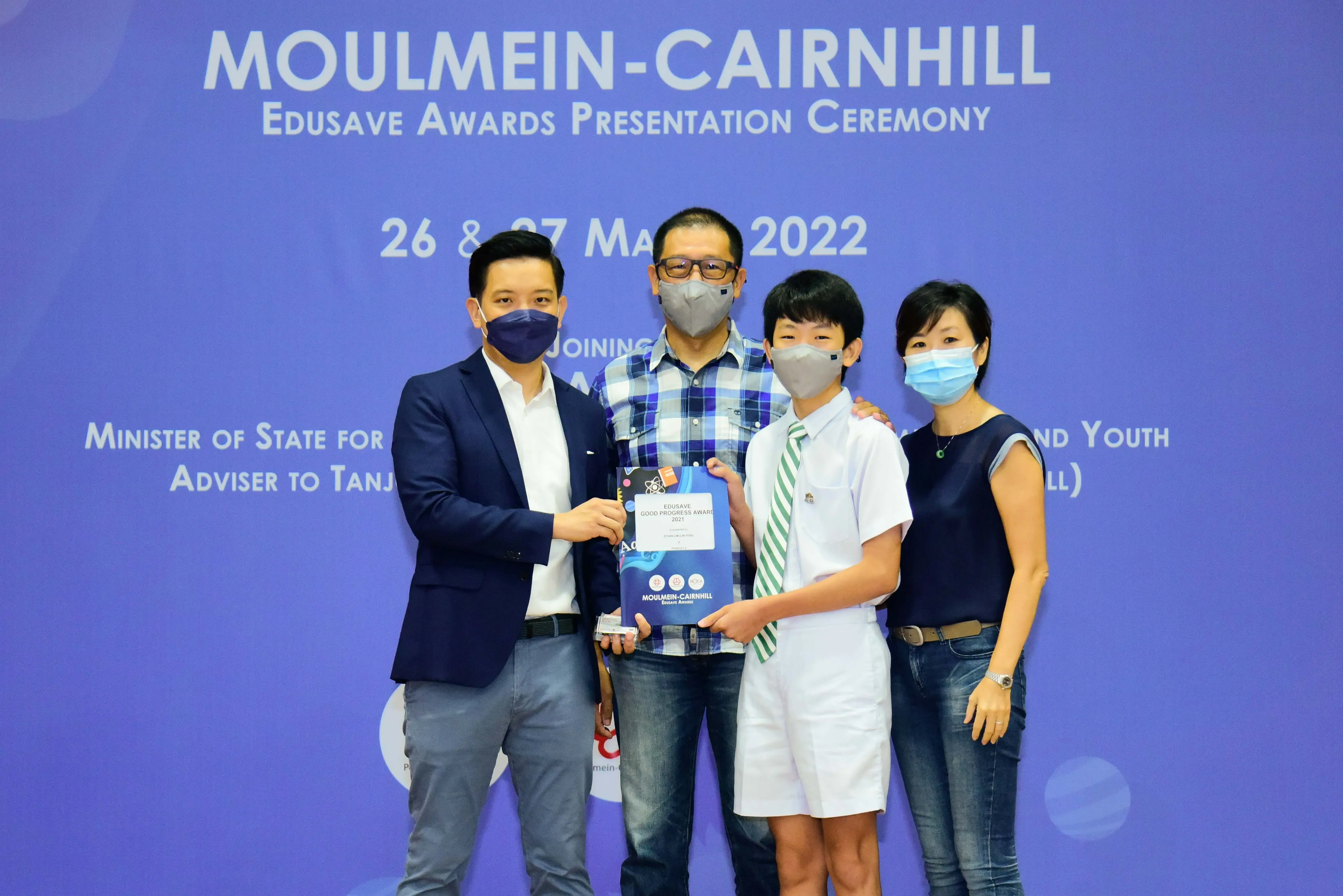 Four masked people pose at the Moulmein-Cairnhill Edusave Awards Ceremony 2022, with a student holding an award.