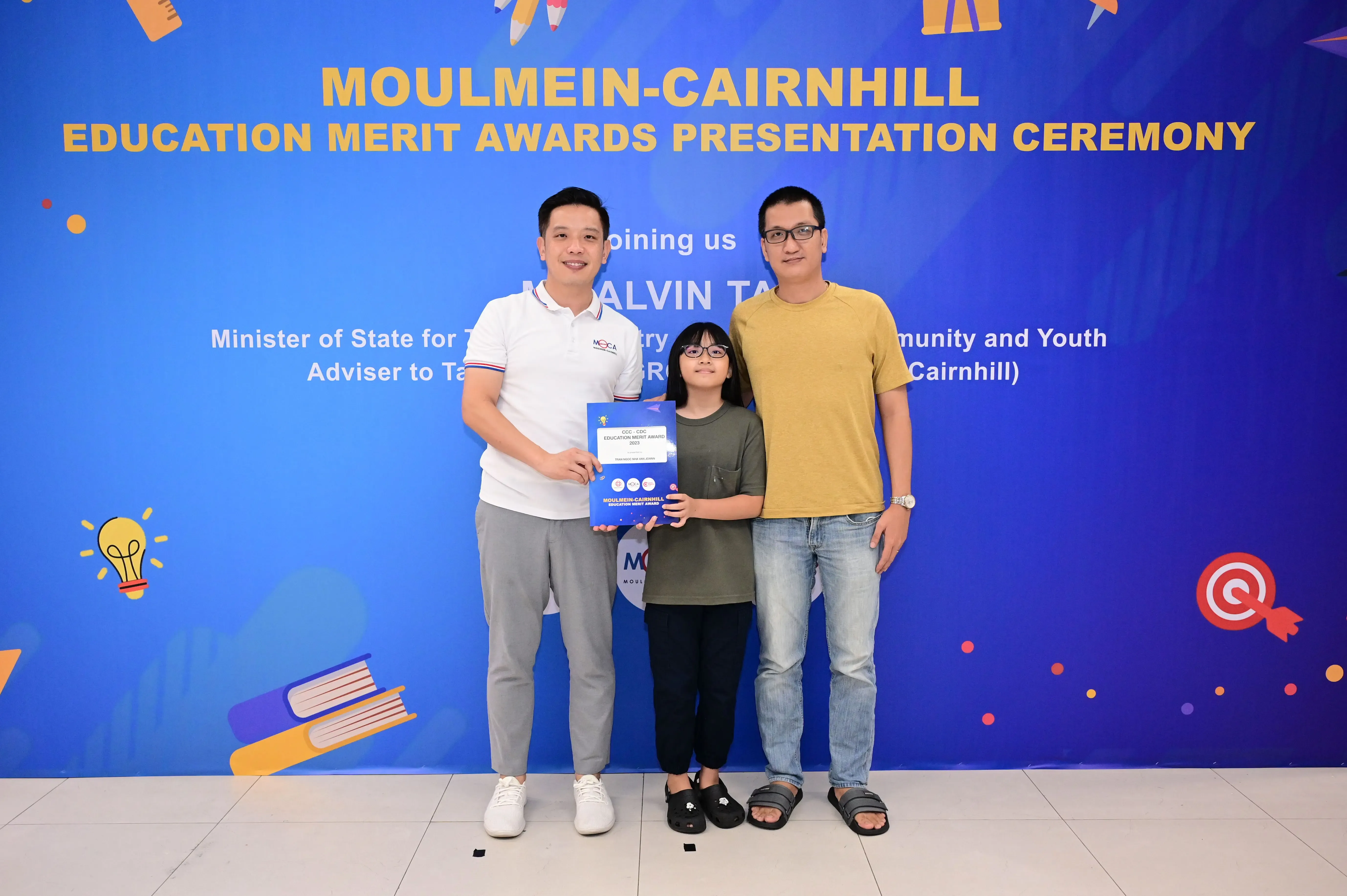 Three people pose before a Moulmein-Cairnhill Education Merit Awards banner, with a girl holding a blue award folder.