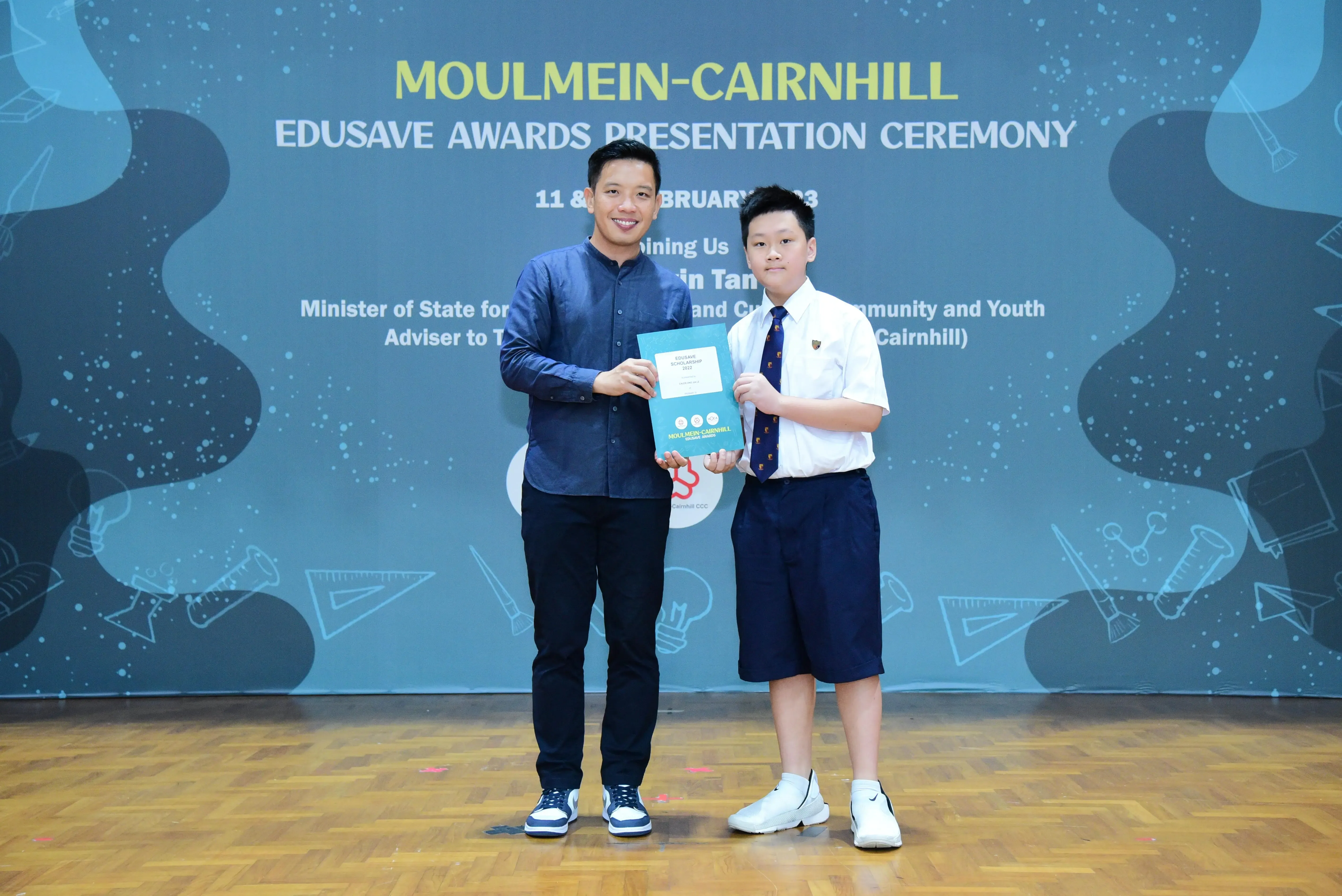 An adult and a young student hold a certificate together at the Moulmein-Cairnhill Edusave Awards Presentation Ceremony.