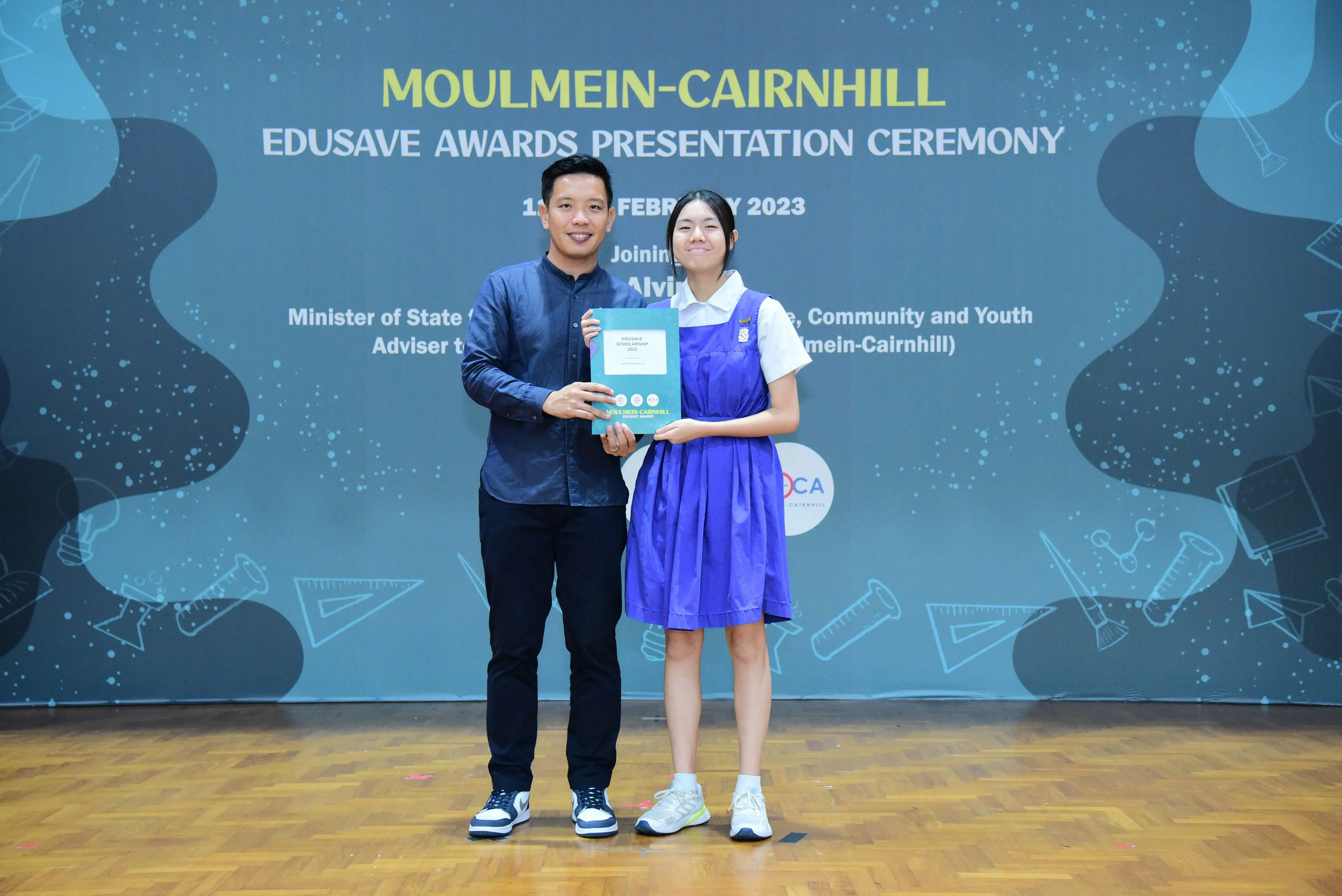 A man and a student in a blue uniform hold a certificate together at the Moulmein-Cairnhill Edusave Awards 2023.