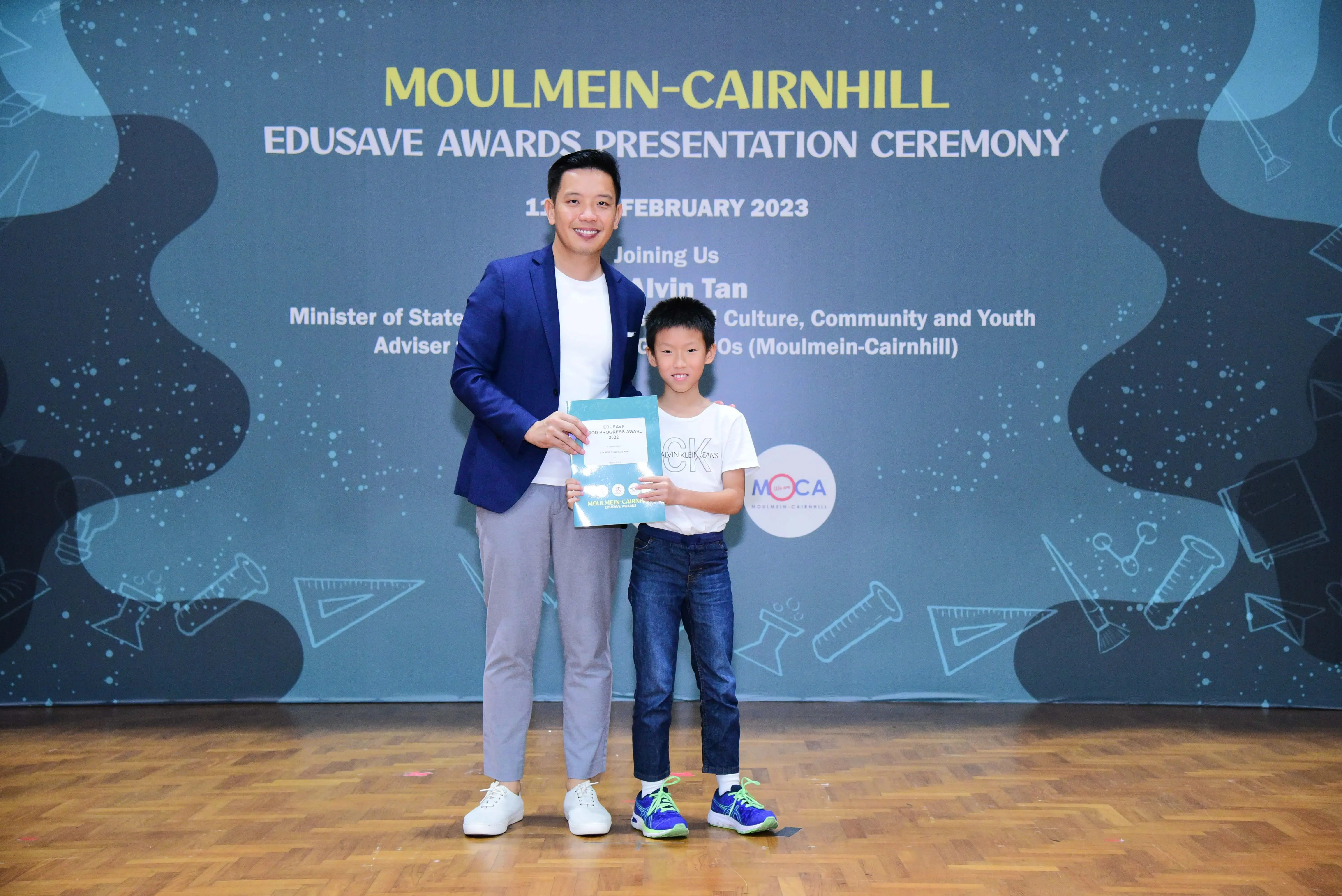 A man in a blue blazer and a young boy hold a certificate at the Moulmein-Cairnhill Edusave Awards Ceremony 2023.