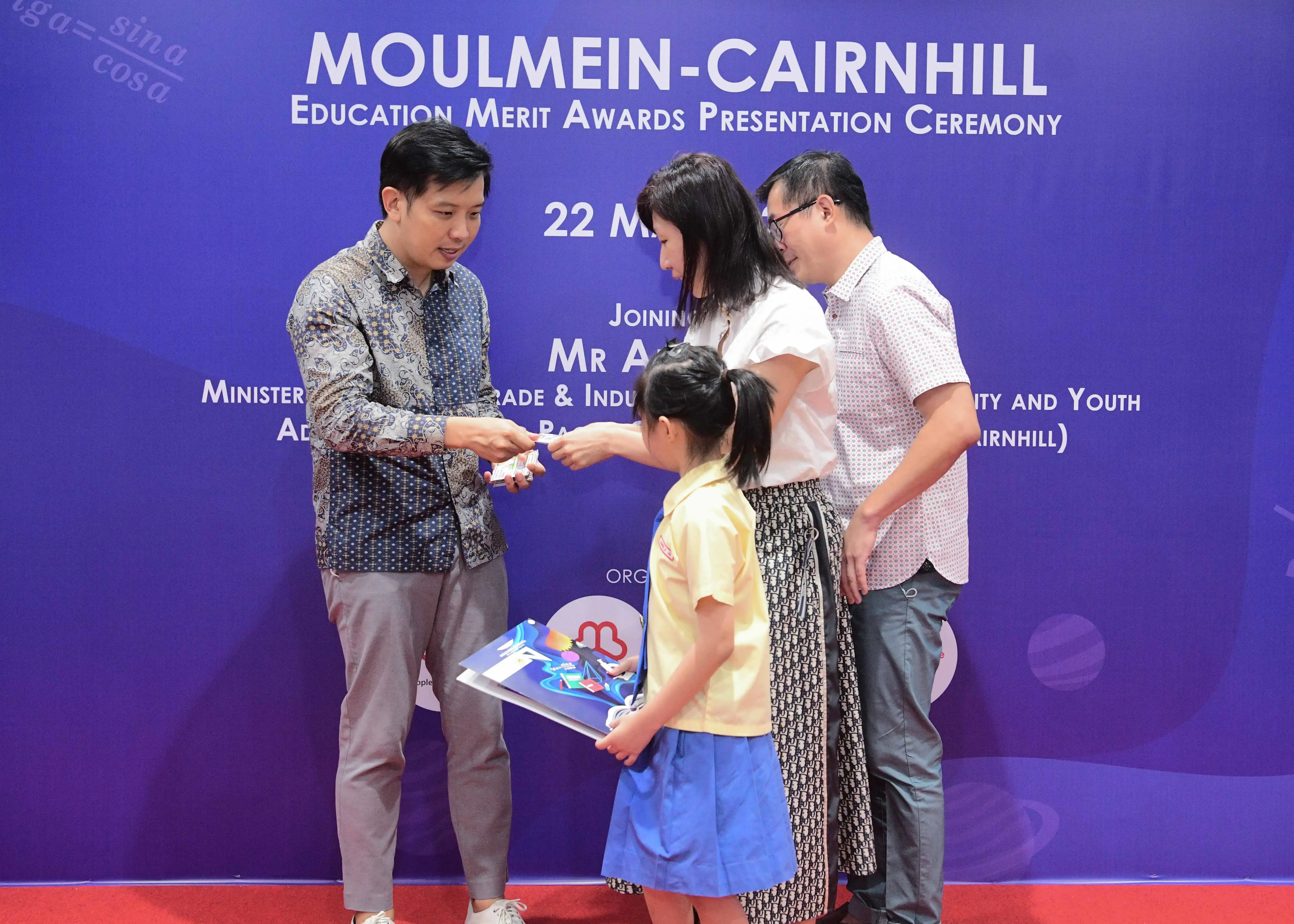 A man presents an award to a young girl in school uniform at the Moulmein-Cairnhill Education Merit Awards Ceremony.