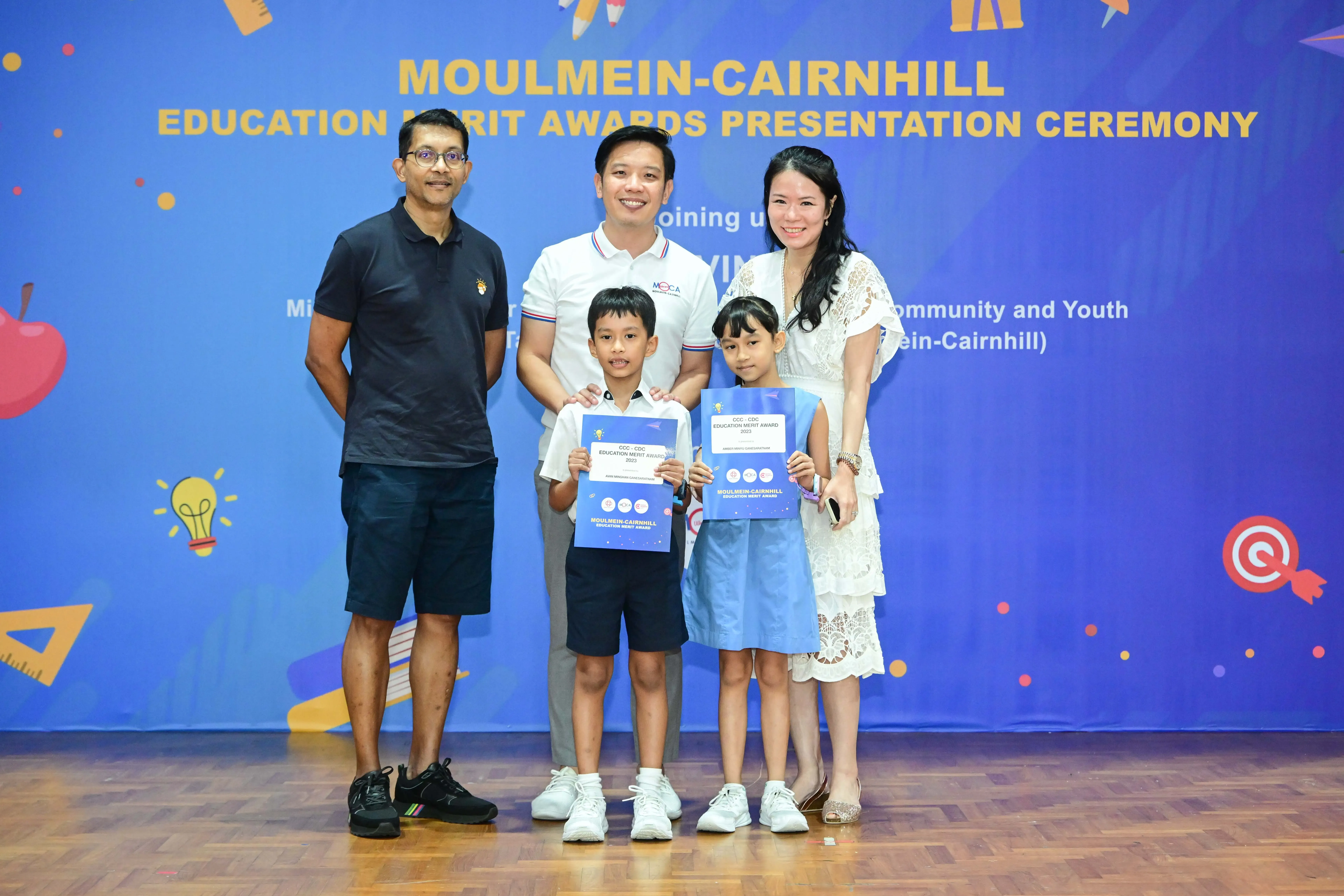 Five people pose before a Moulmein-Cairnhill Education Merit Awards banner, two children holding award certificates.