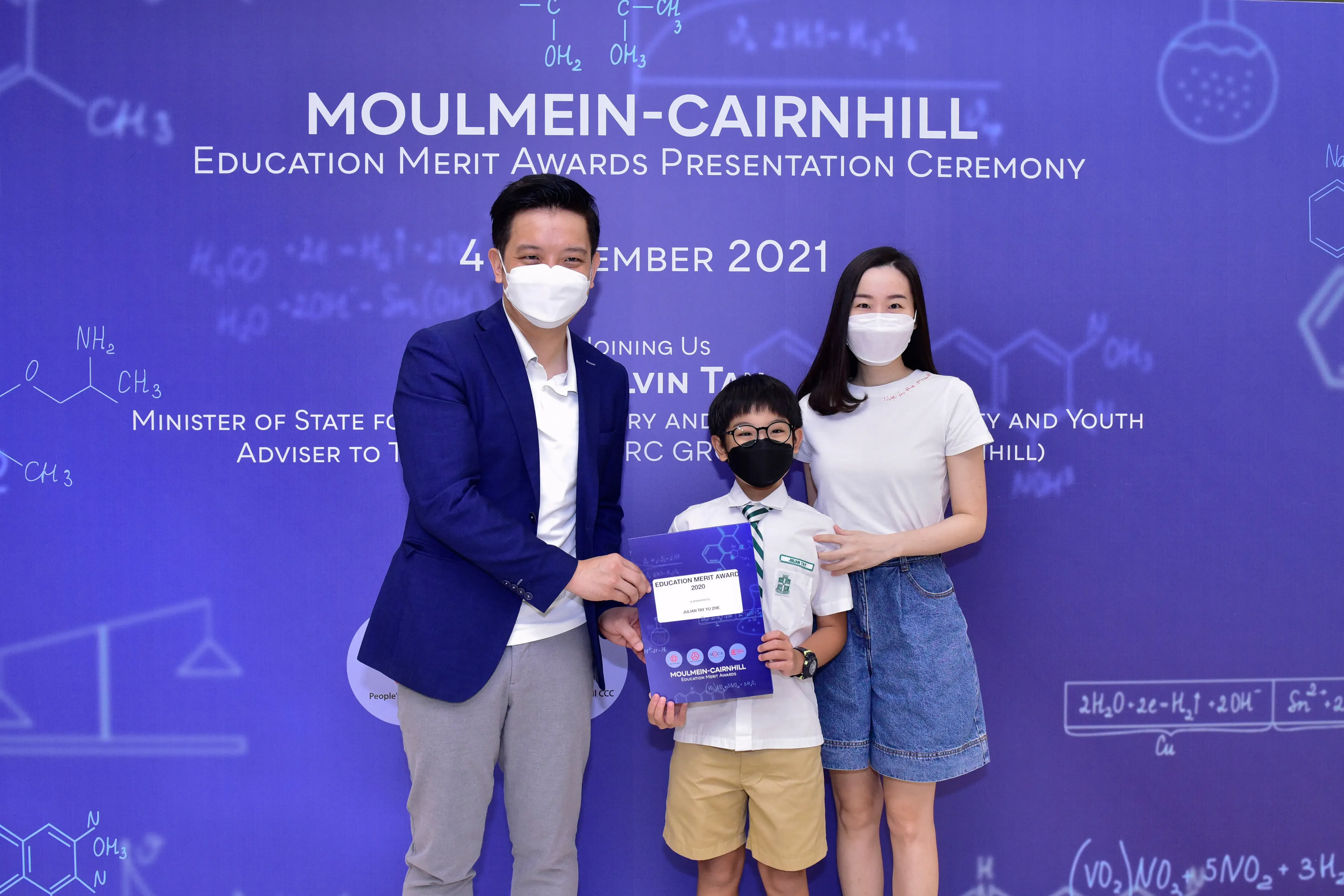 Three masked people pose at the Moulmein-Cairnhill Education Merit Awards 2021, with a boy holding a certificate.